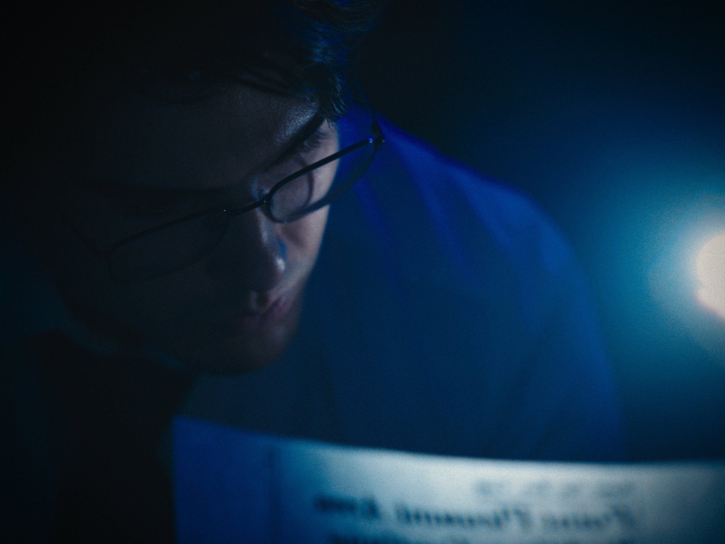 A person wearing glasses is looking down in a dark room with a blue light illuminating part of their face.