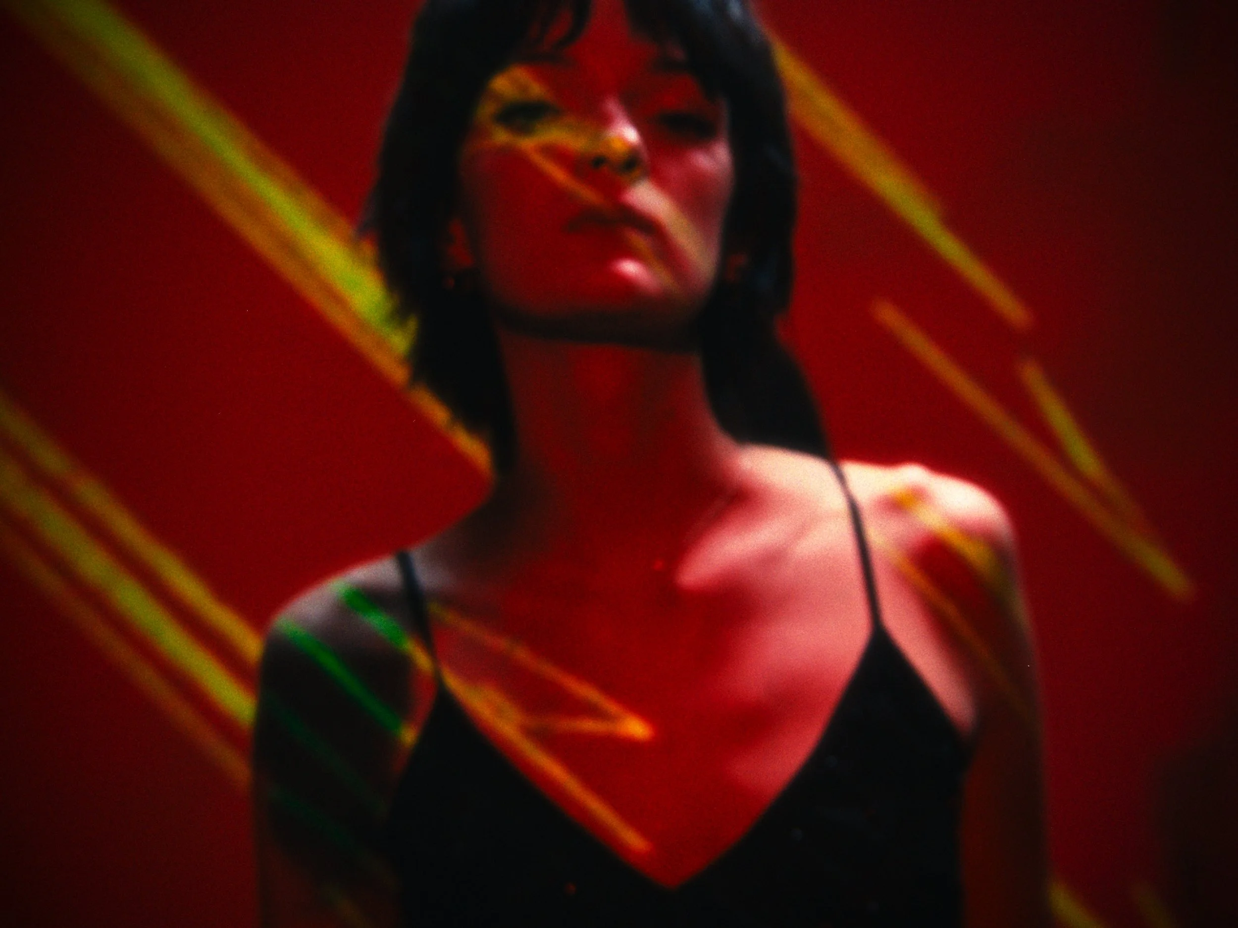A woman with black hair wearing a black spaghetti strap top, standing against a red background with yellow and green streaks of light.
