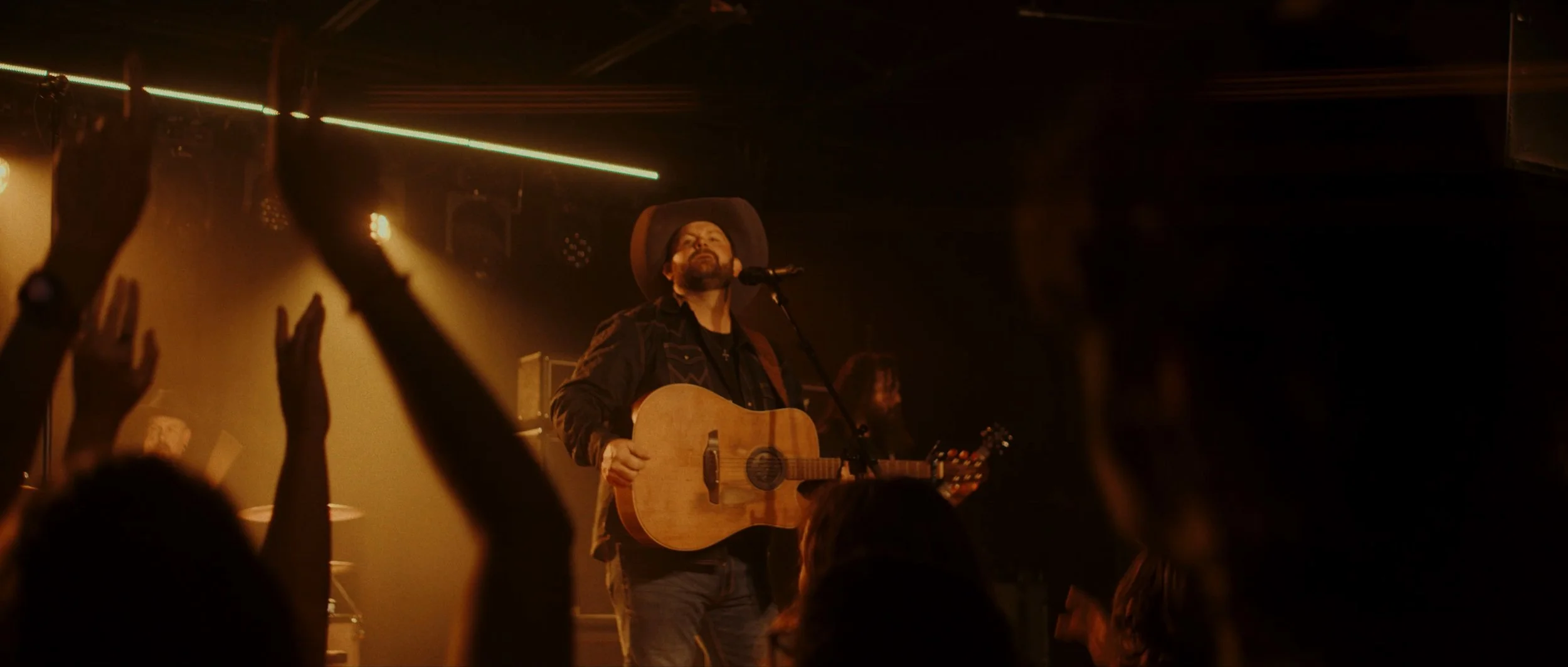 Musician performing on stage with a guitar, wearing a cowboy hat, with audience raising hands in front.