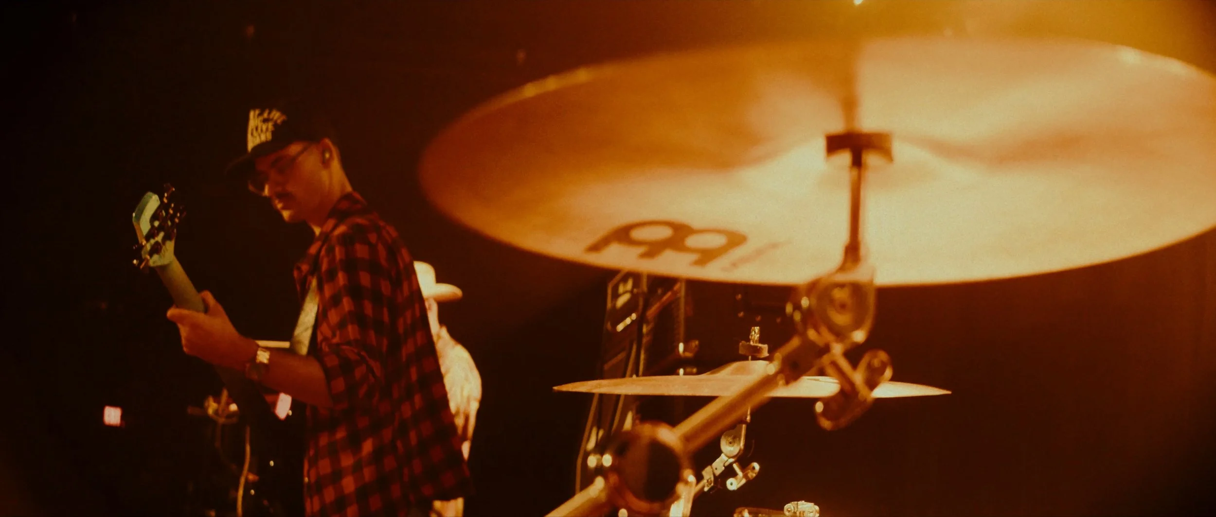 A person wearing a red and black checkered shirt and a baseball cap, playing an electric guitar, with a drum set visible in the foreground.