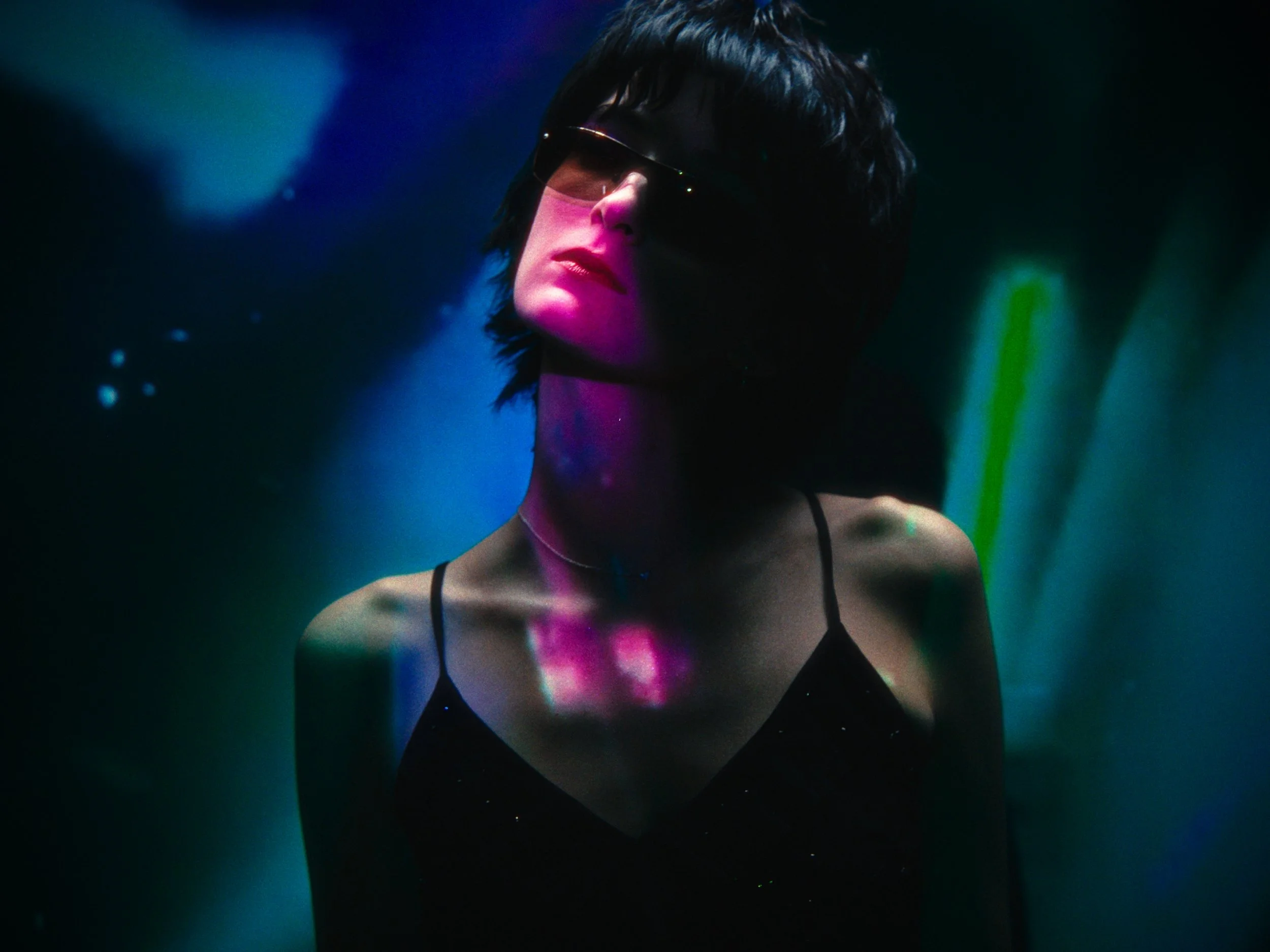 A woman with short black hair wearing sunglasses and a black spaghetti strap top in a dark, colorful environment with neon lighting.
