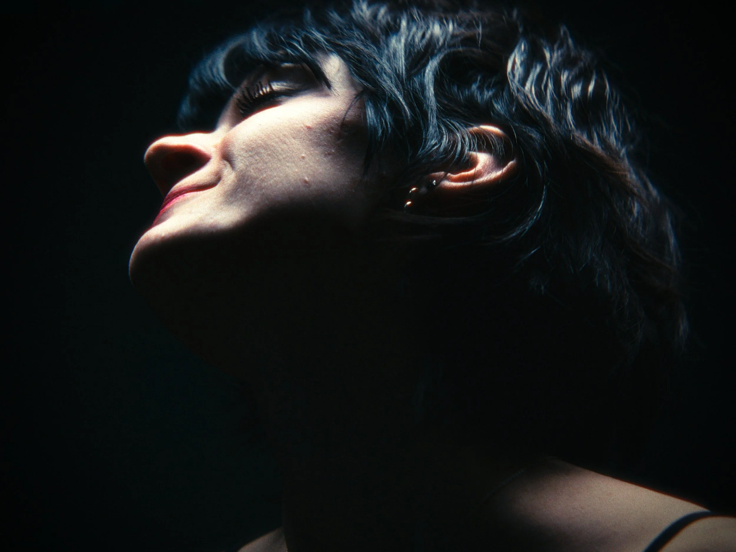 Close-up side profile of a woman with short dark hair against a black background, illuminated softly.