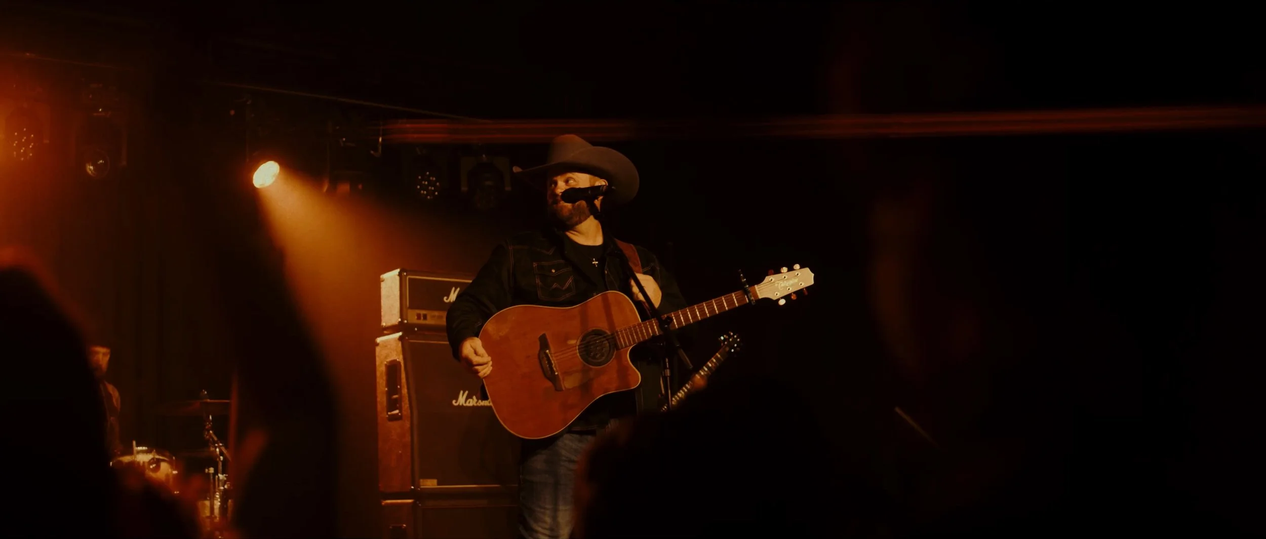 A male musician wearing a cowboy hat plays an acoustic guitar on stage, illuminated by warm orange stage lights, with a drum set and Marshall amplifier in the background.