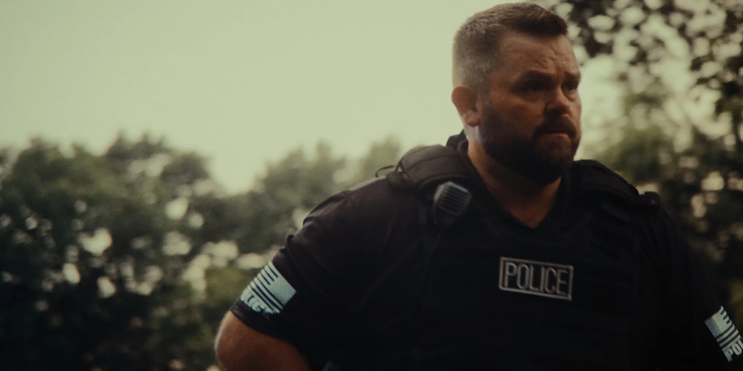 A police officer with a beard and short hair, wearing a black uniform with police patches on sleeves, is outdoors with blurred trees in the background.