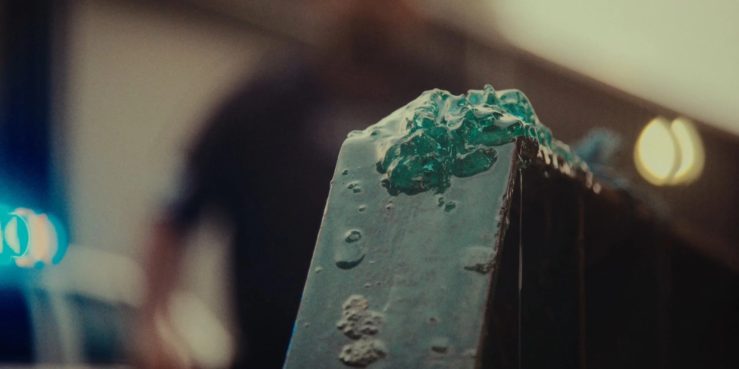 Close-up of a metal spatula with melted greenish-blue wax or resin on it, in a dimly lit environment.