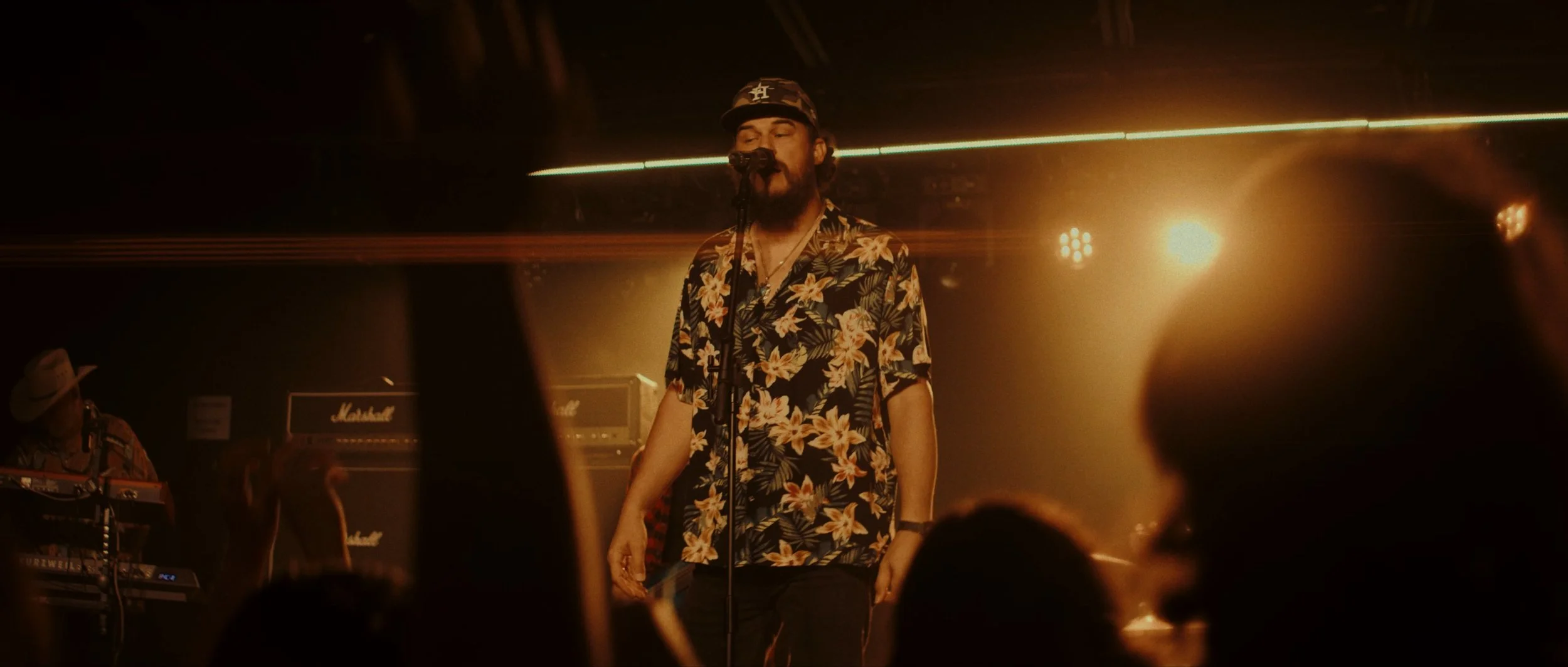 A man with a beard and sunglasses performing on stage, wearing a floral shirt and a cap. There is a musician in a cowboy hat playing keyboard in the background, with music equipment and amplifiers around him.
