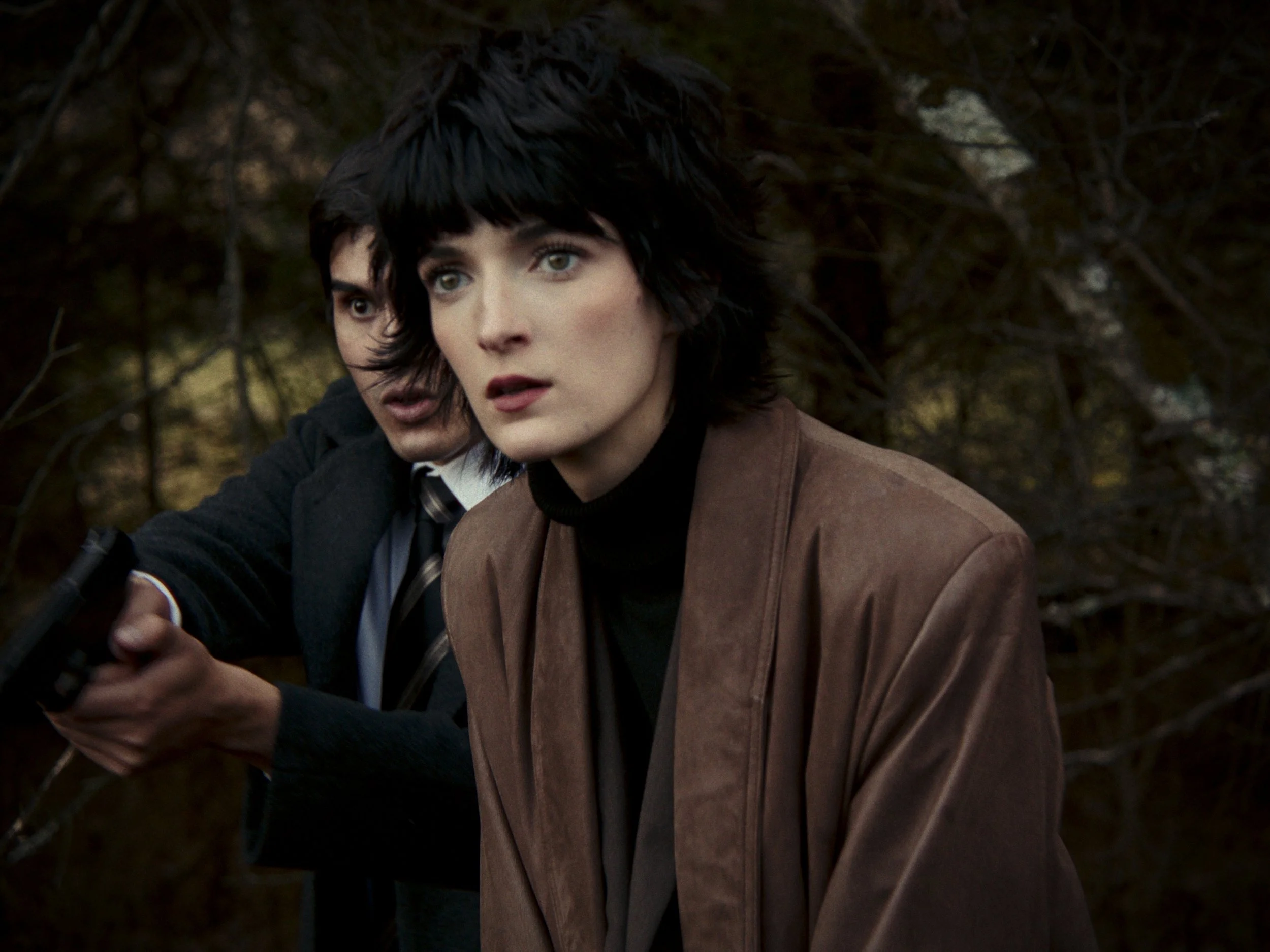 Woman with short black hair and brown jacket appears tense in a wooded area, while a man behind her points a gun. X- files
