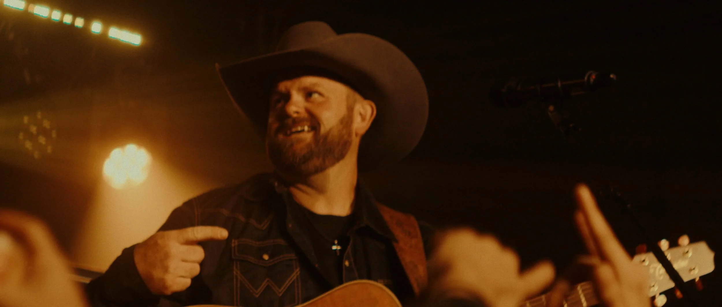 A man with a beard and a cowboy hat smiling while playing a guitar in a dimly lit setting, with audience members' hands raised.