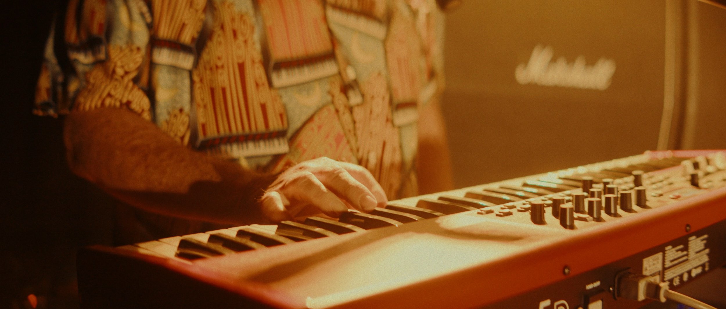 Close-up of a person playing a red keyboard with their right hand, wearing a colorful, patterned long-sleeve shirt, in a warm lighting environment.