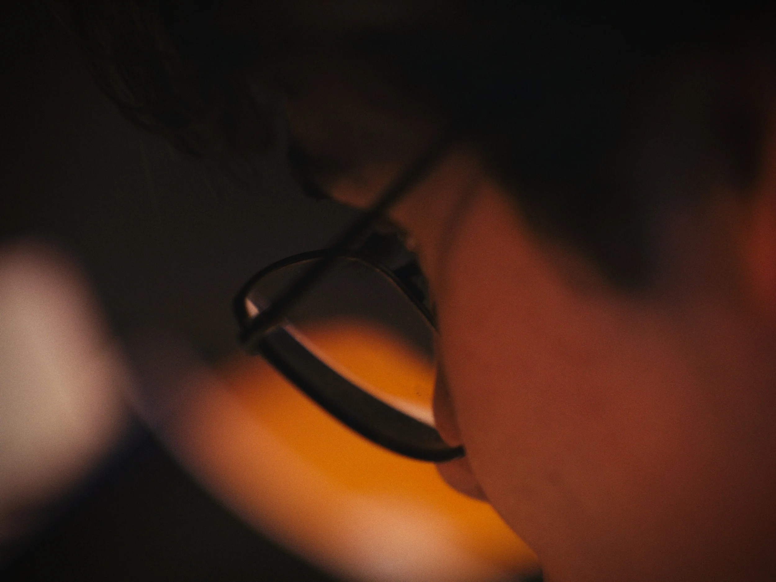 Close-up of a person's face with glasses, focusing on their eye and nose with warm orange lighting.