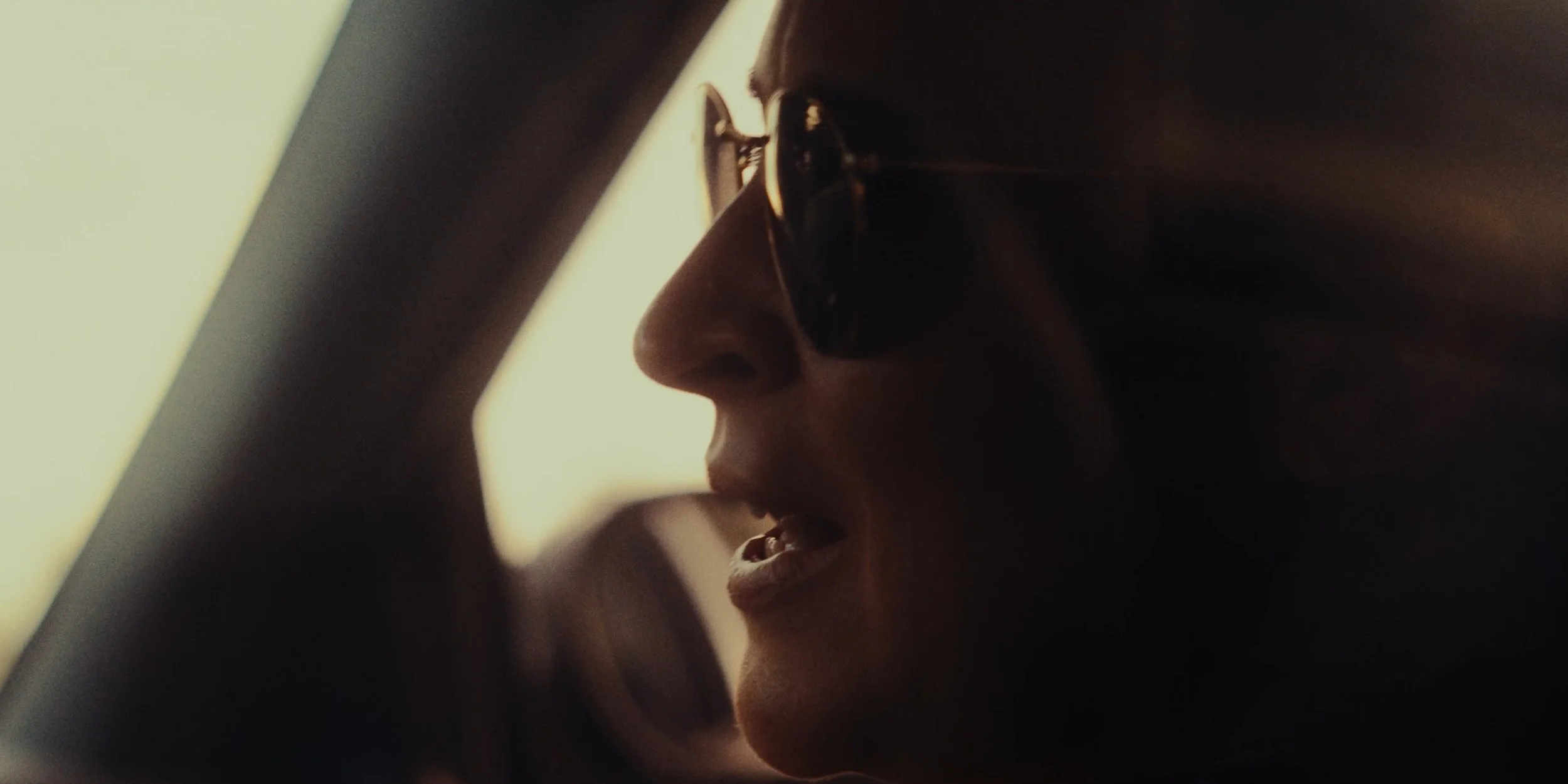Close-up of a woman wearing sunglasses, speaking or singing, with side profile view, in a dimly lit setting.