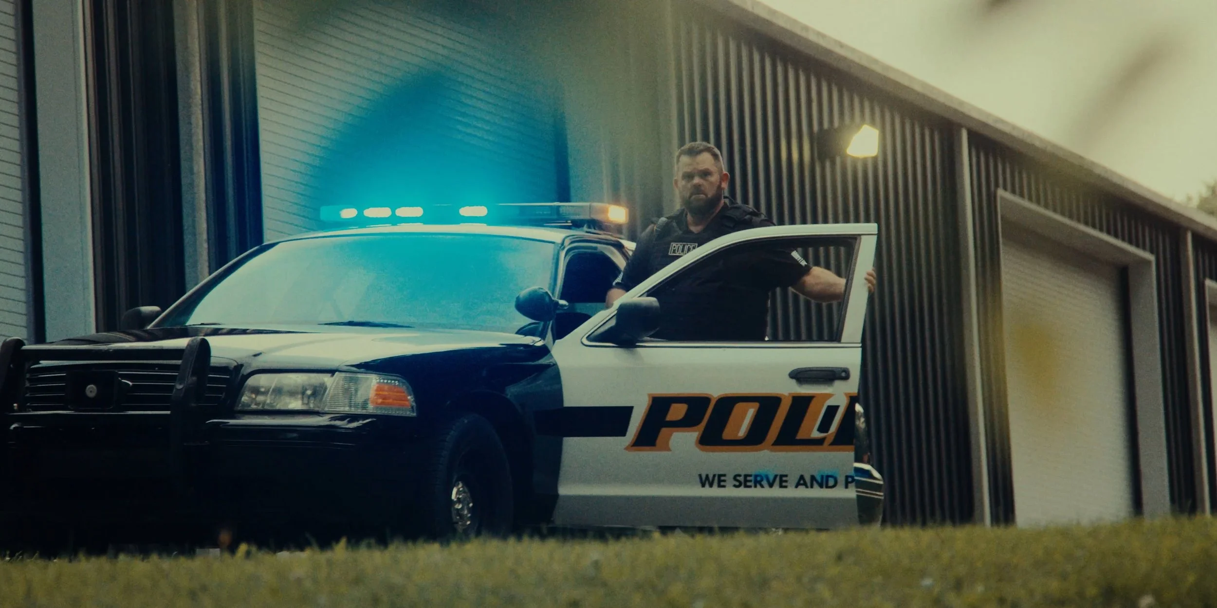 A police officer standing next to an open police car door outside a modern building with garage doors and exterior lights.