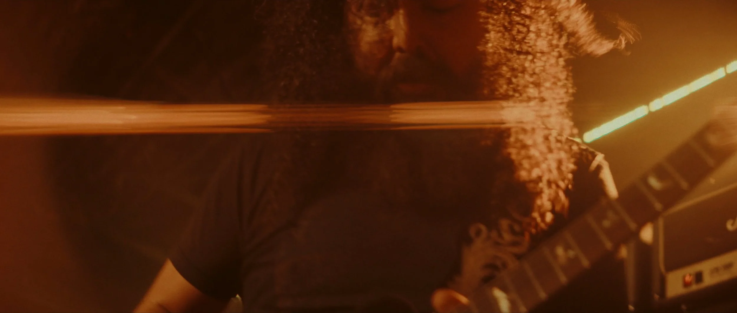 A person with long curly hair playing an electric guitar in a dimly lit environment with orange stage lighting.
