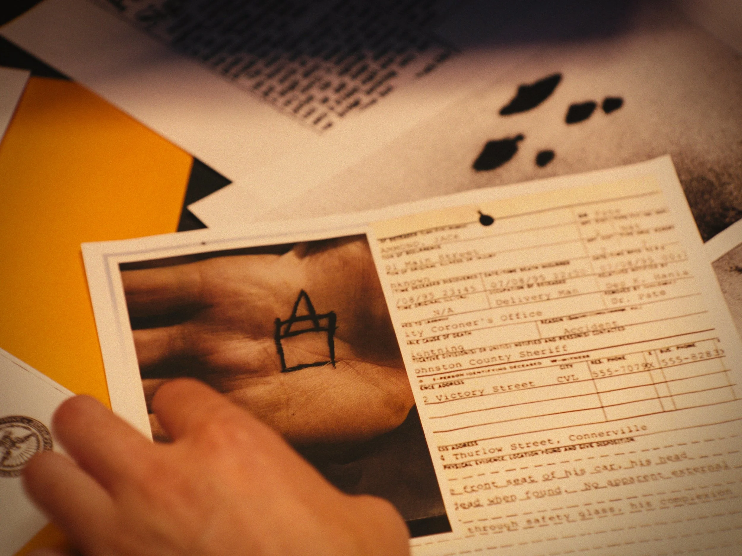 Close-up of a hand holding a photograph of a hand with a black marker drawing of a house on it, alongside a printed document with handwritten and typed text. Items are on a cluttered table with paper and a yellow folder.