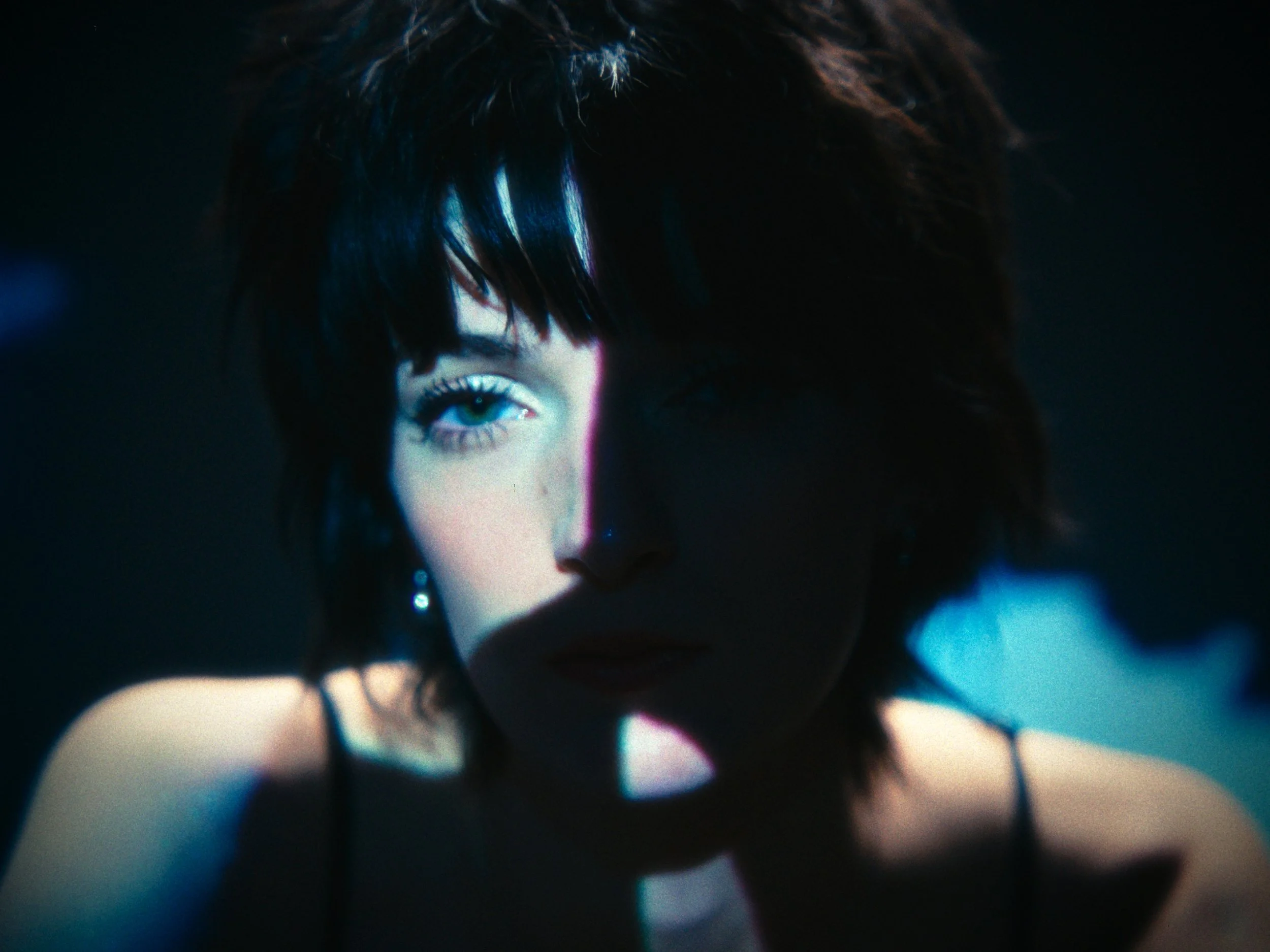 Close-up of a woman with dark hair, one eye visible, illuminated with colorful lighting in a dark setting.