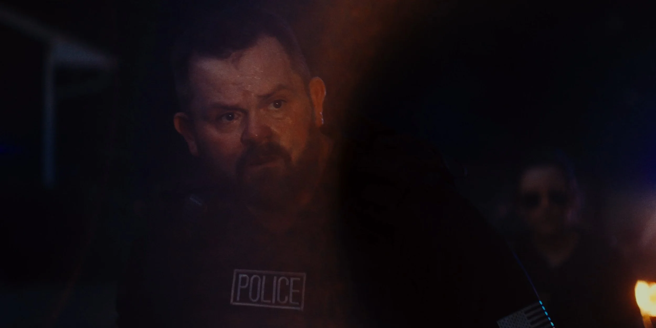A police officer with a beard and short hair, wearing a uniform with a badge that says 'POLICE,' is looking intently to his left in a dark setting.