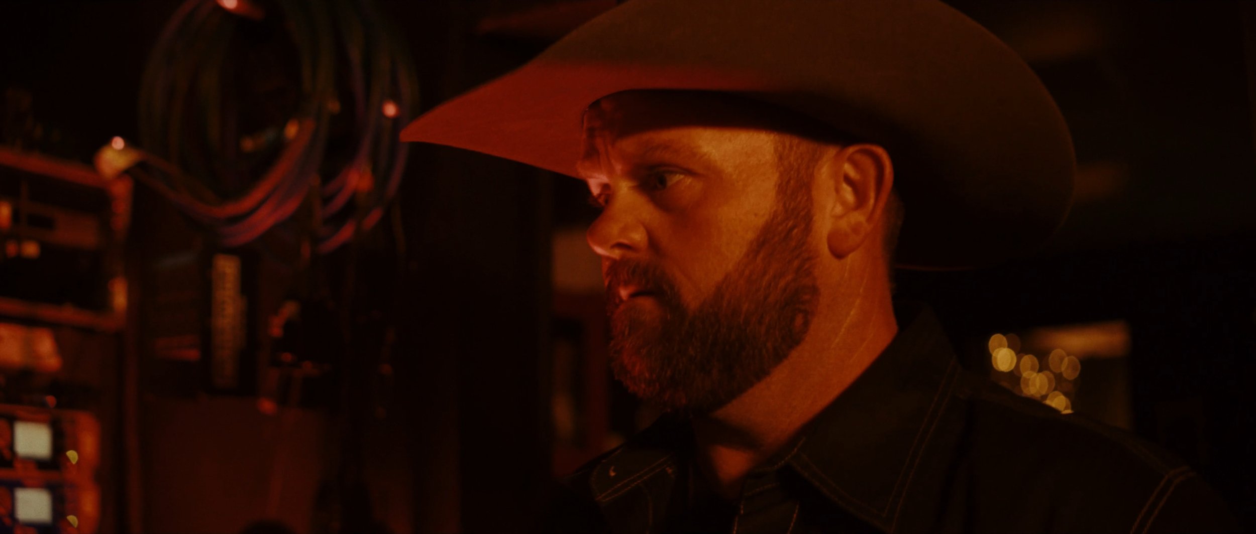 A man wearing a cowboy hat with a beard and mustache, in a dimly lit environment with red lighting.