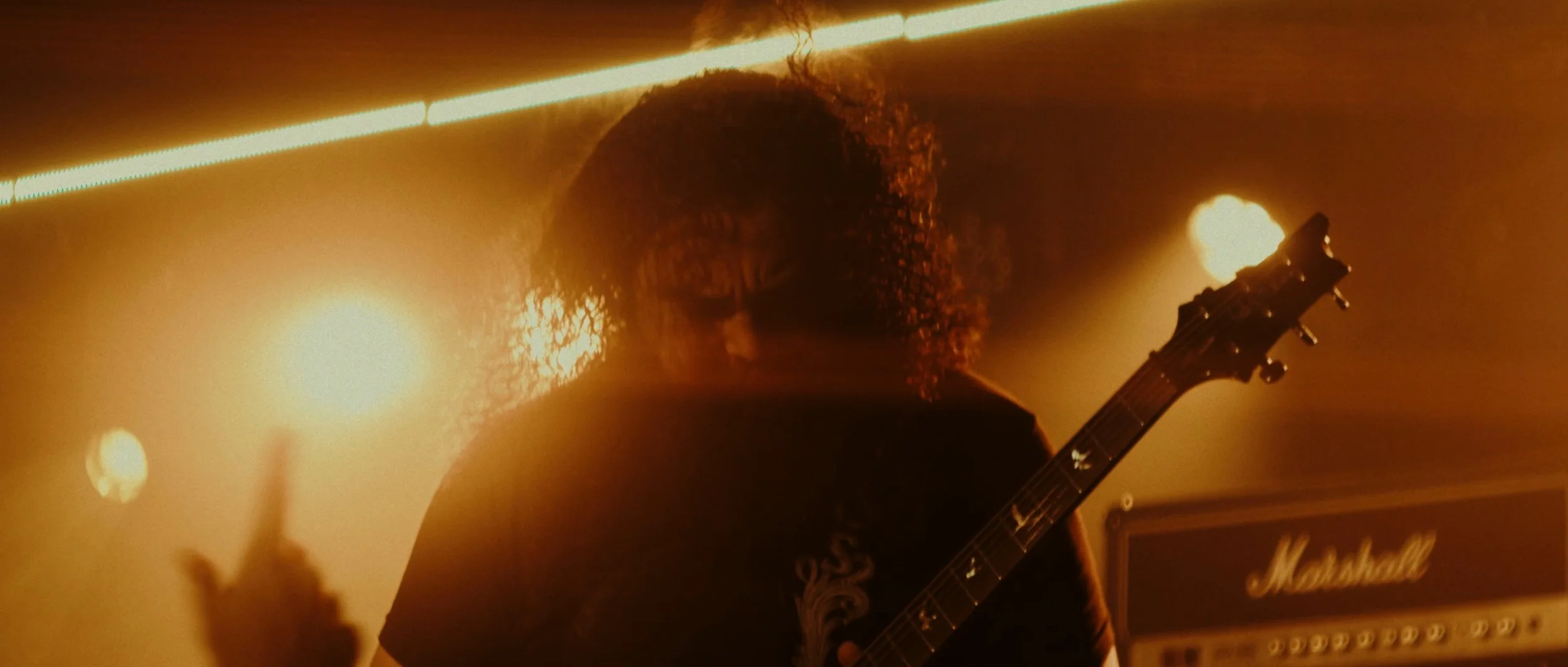 A musician with curly hair playing an electric guitar on stage, illuminated by warm orange stage lights, with a Marshall amplifier visible in the background.