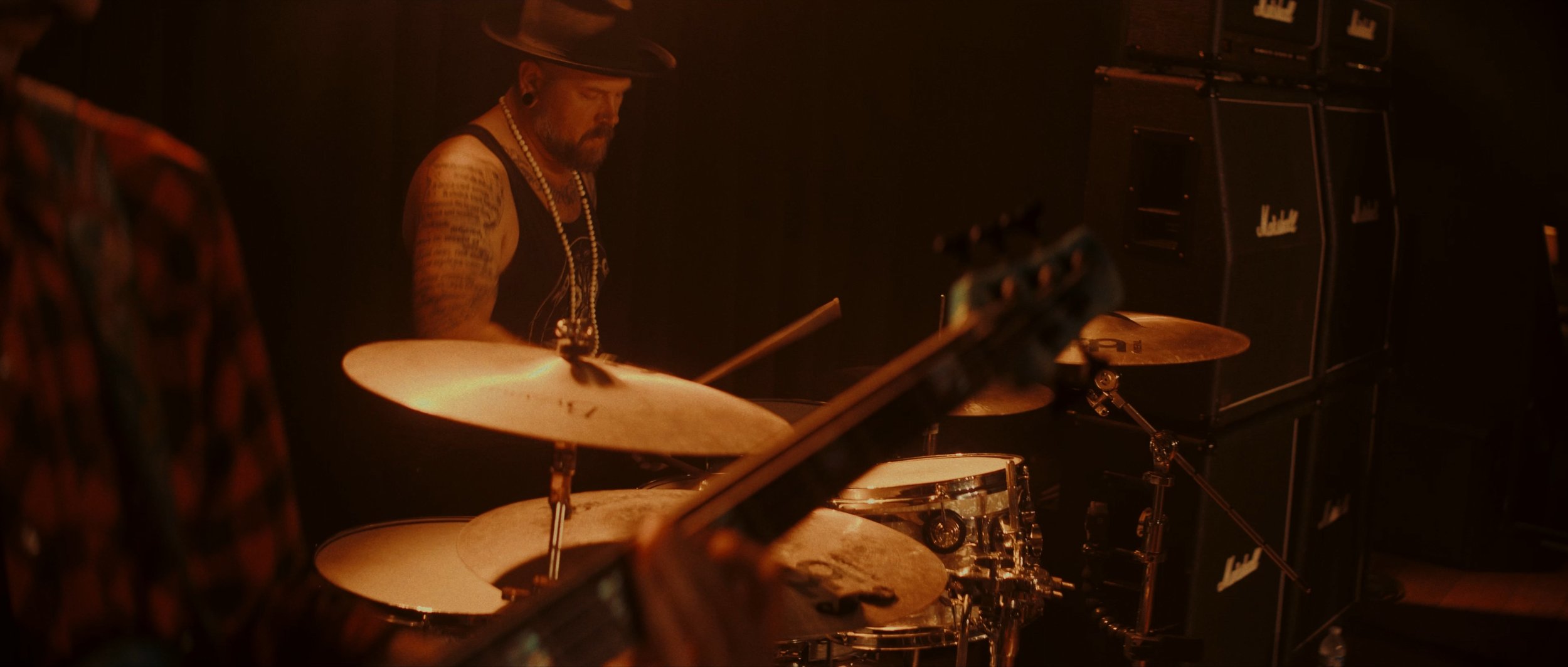 A musician playing drums on stage, with Marshall amplifier stacks visible in the background, in a dark, warm-lit setting.
