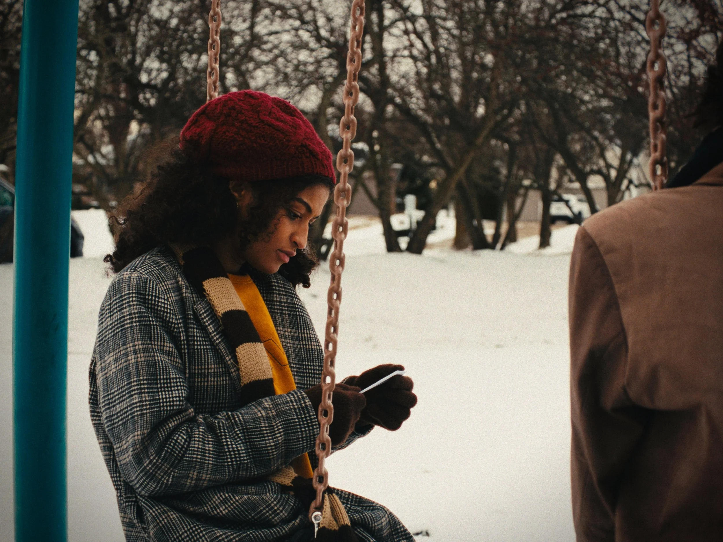 A woman with curly hair wearing a red knit hat and checkered coat sitting on a swing in a snowy park, looking at her phone. Another person is partially visible on the right, wearing a brown jacket.