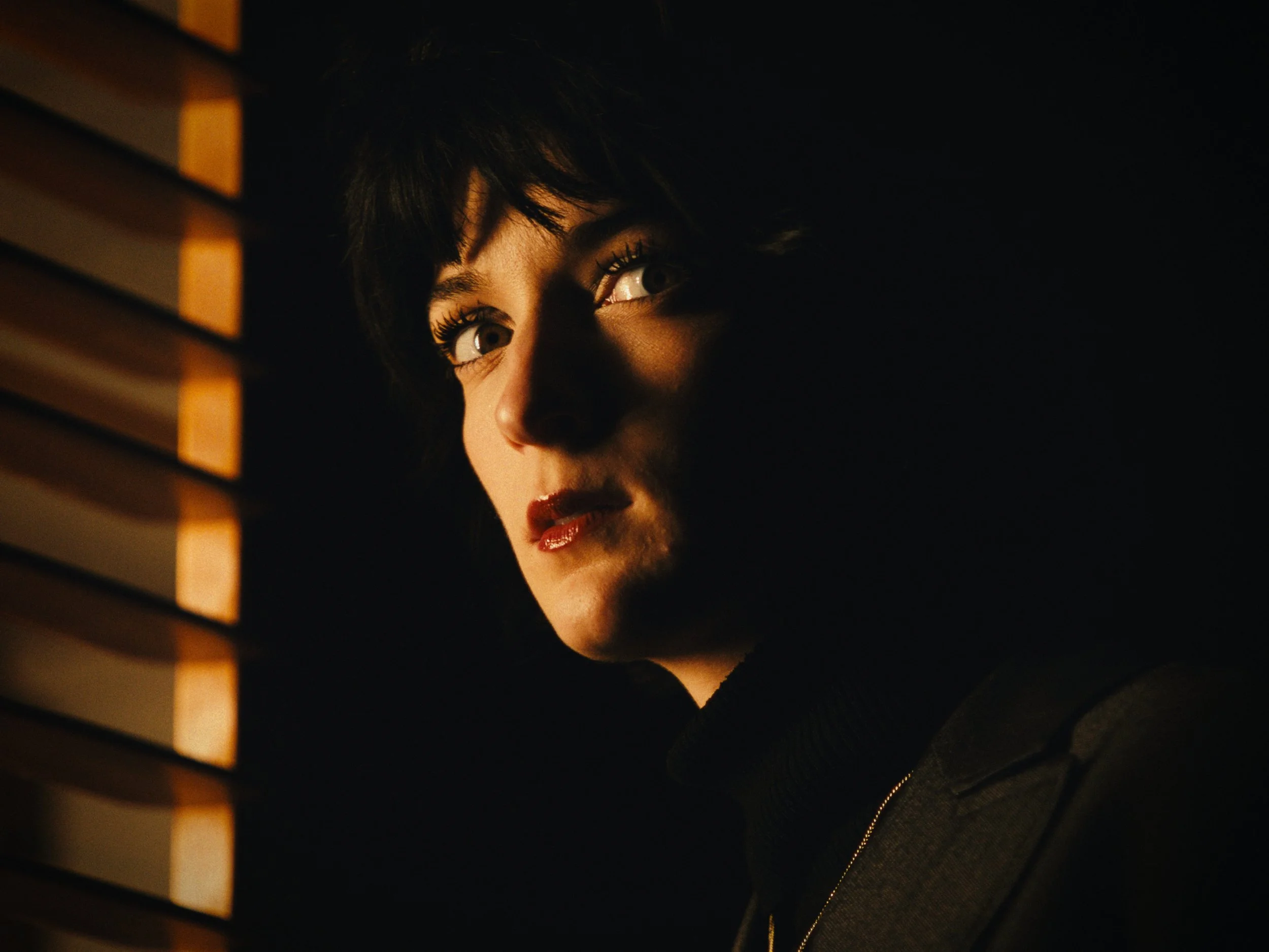A woman with short dark hair and makeup looking through window blinds with warm light illuminating her face.