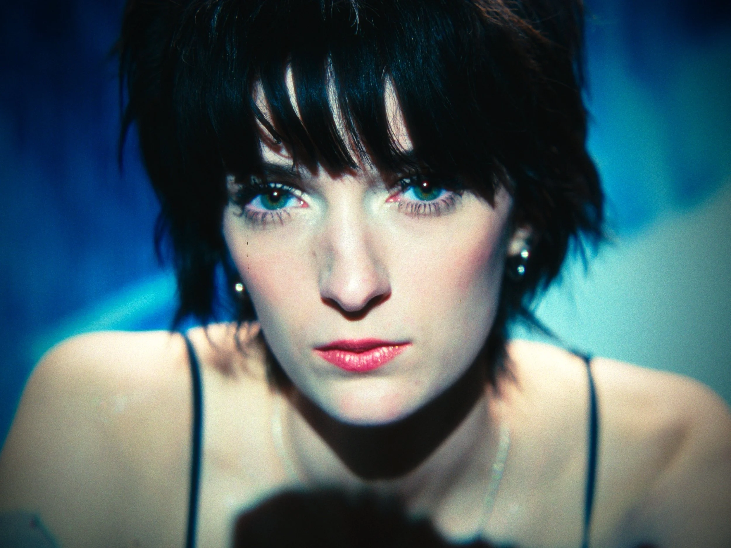 Close-up of a woman with short dark hair and blue eyes, wearing earrings and a black spaghetti strap top, looking directly at the camera.