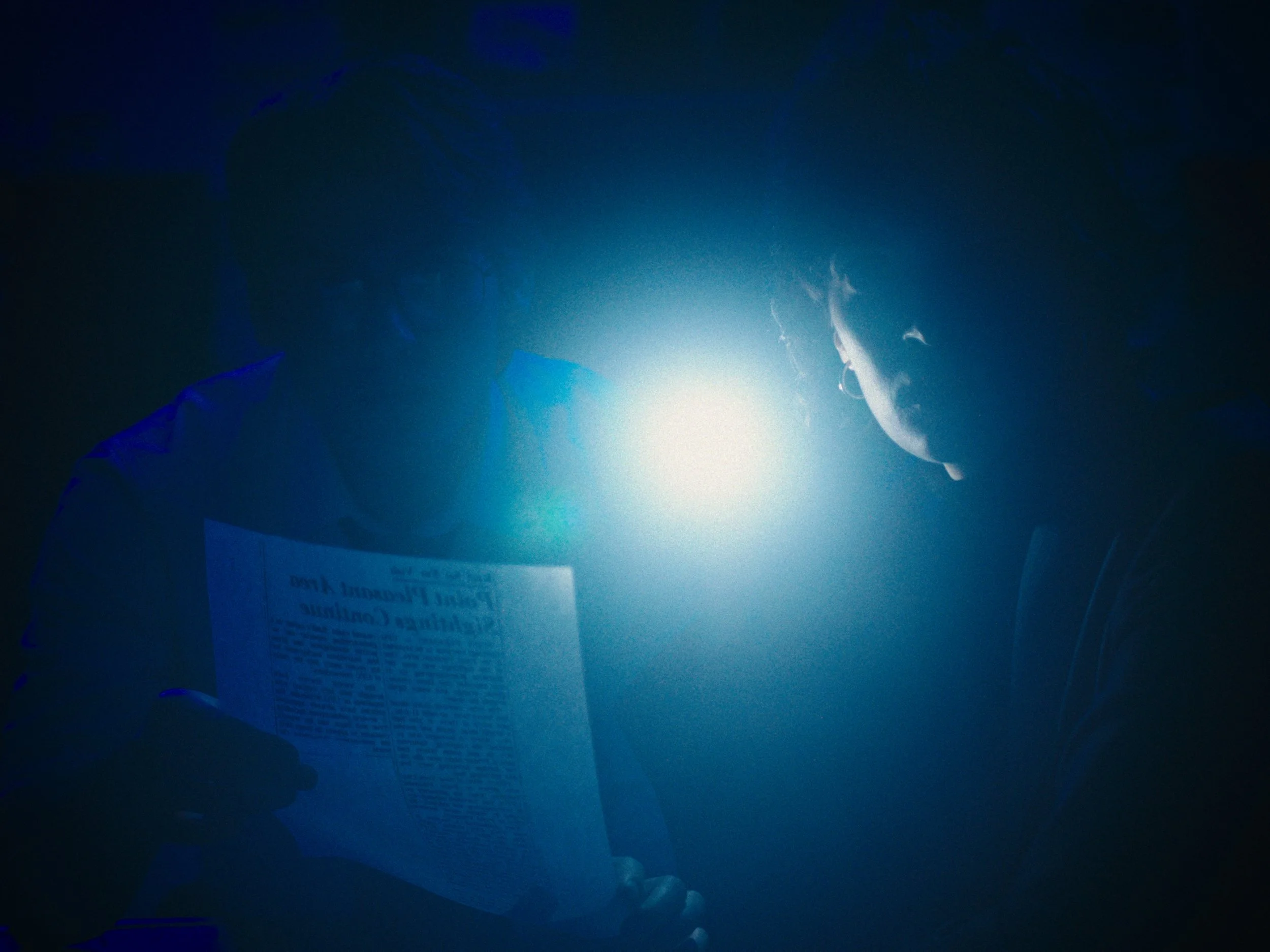 Two people in darkness, illuminated by bright blue light, reading a newspaper.
