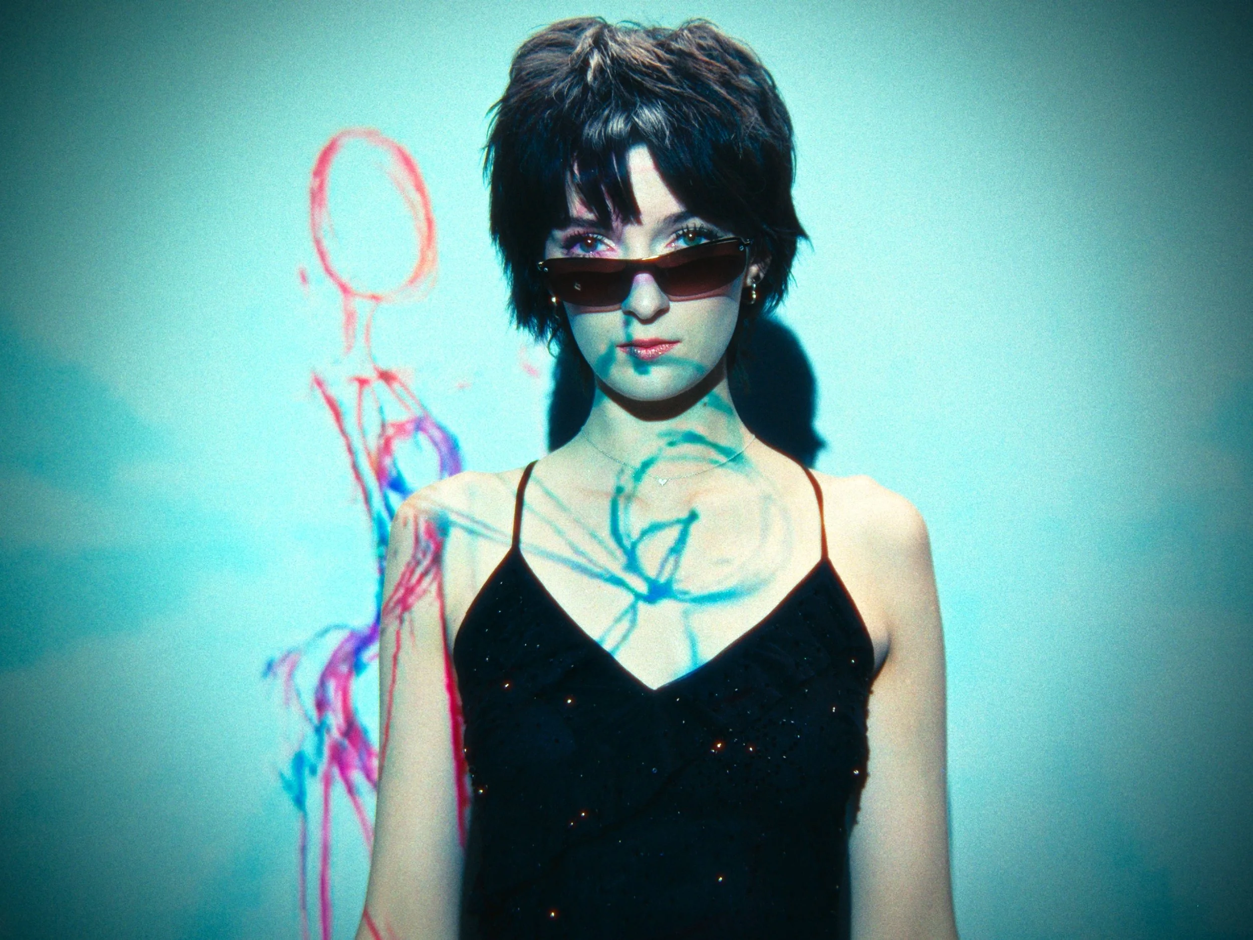 A woman with short dark hair wearing sunglasses and a black dress, standing in front of a wall with colorful abstract drawings projected behind her.