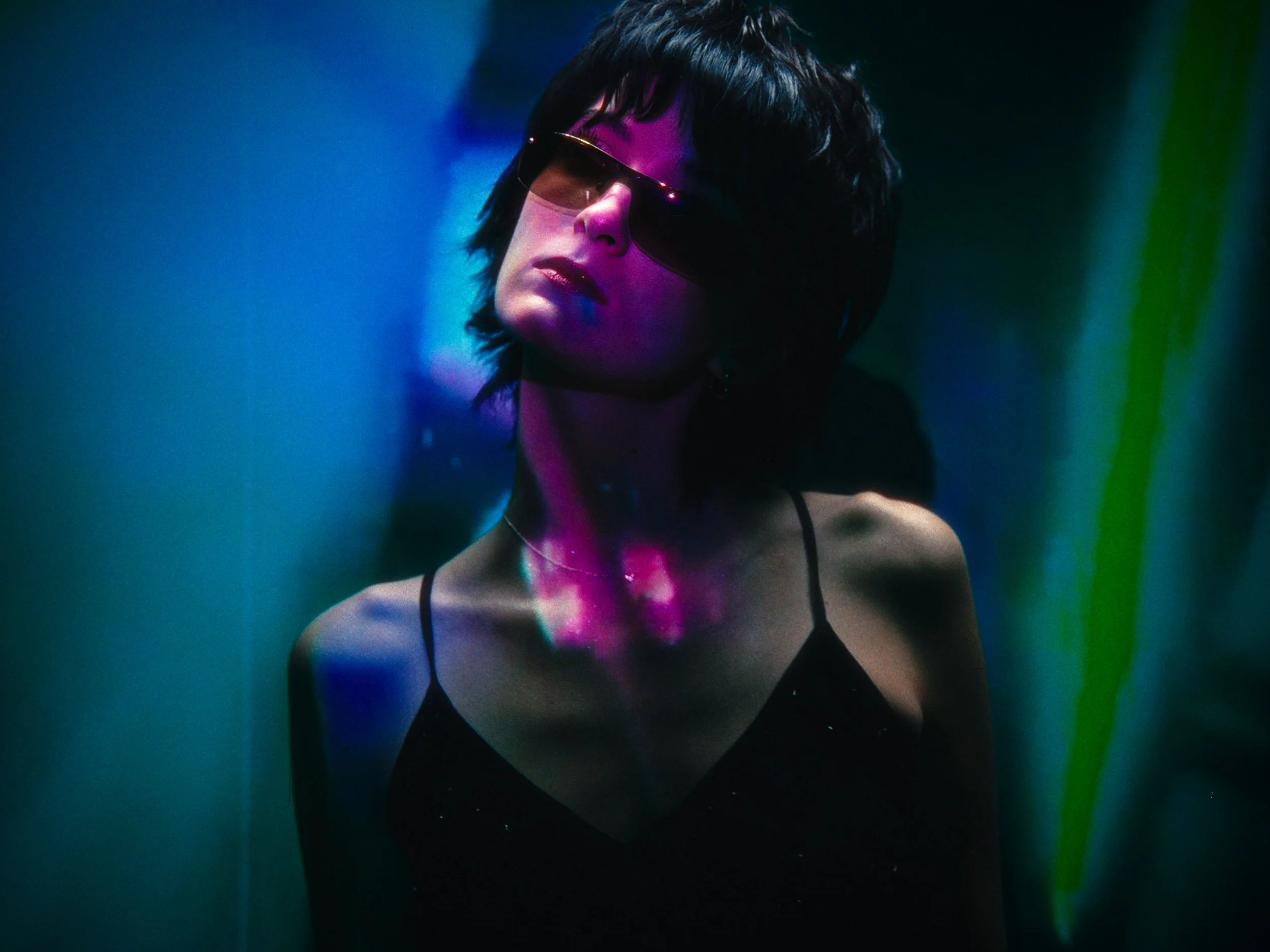A woman with short black hair wearing sunglasses and a black spaghetti strap top, illuminated by colorful neon lights.