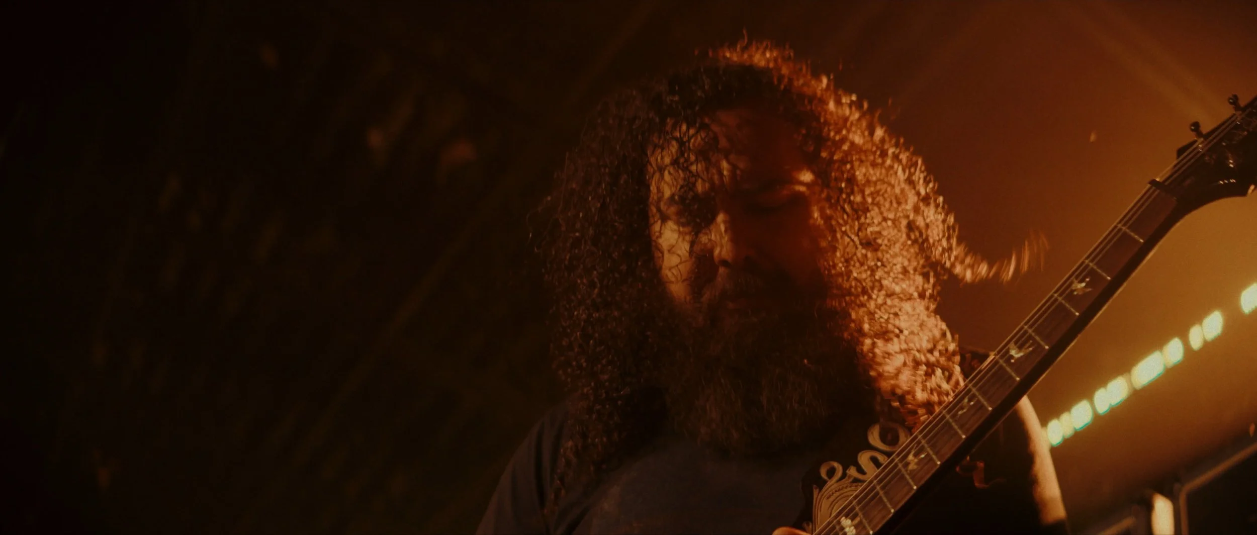 A man with long curly hair and a beard playing an electric guitar in a dimly lit, fiery orange setting.