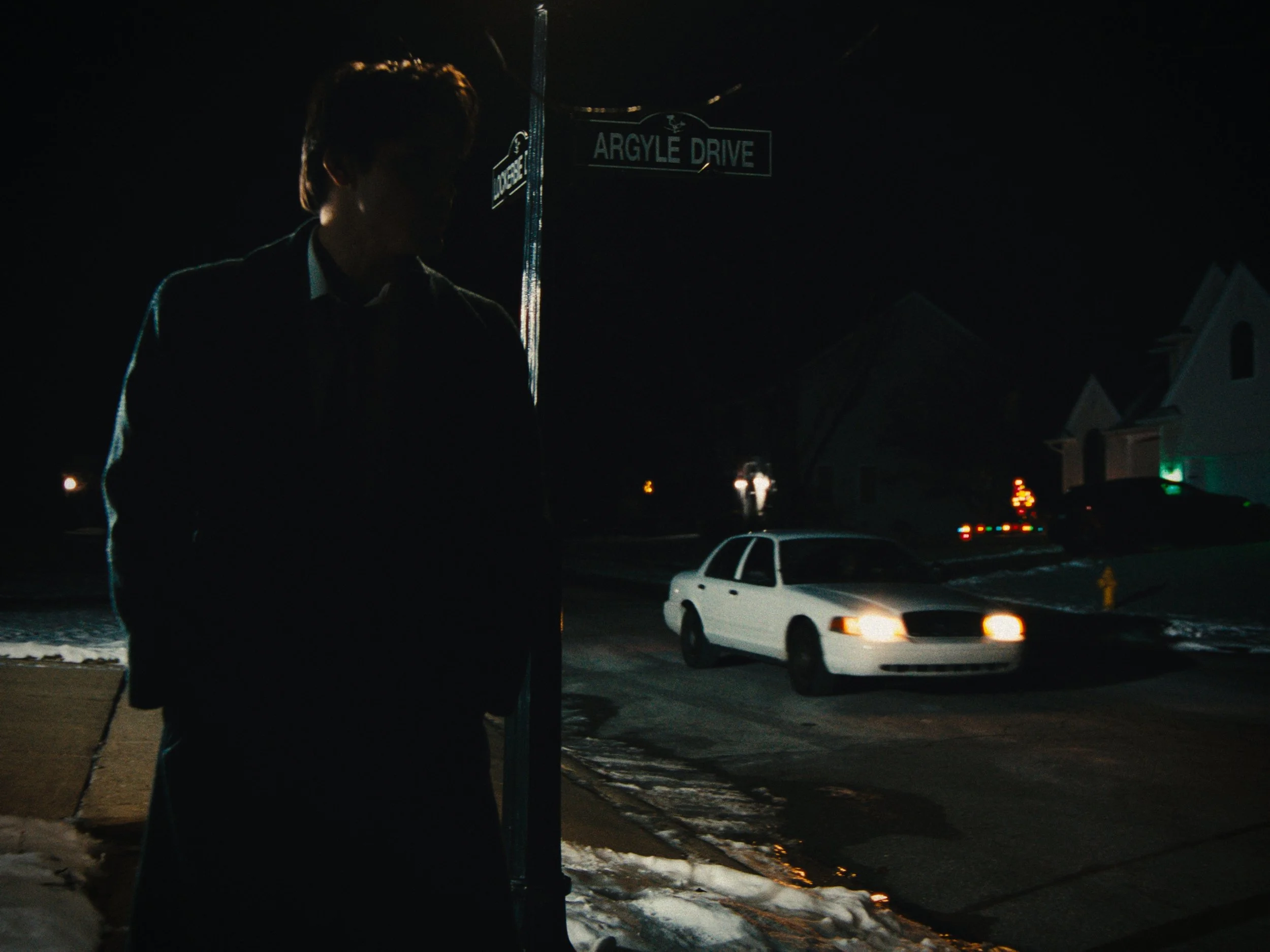 A person standing at a street corner at night, silhouette visible against the dark background. A car with headlights on is passing by on the street. The street sign reads "Argyle Drive." Snow is on the ground.