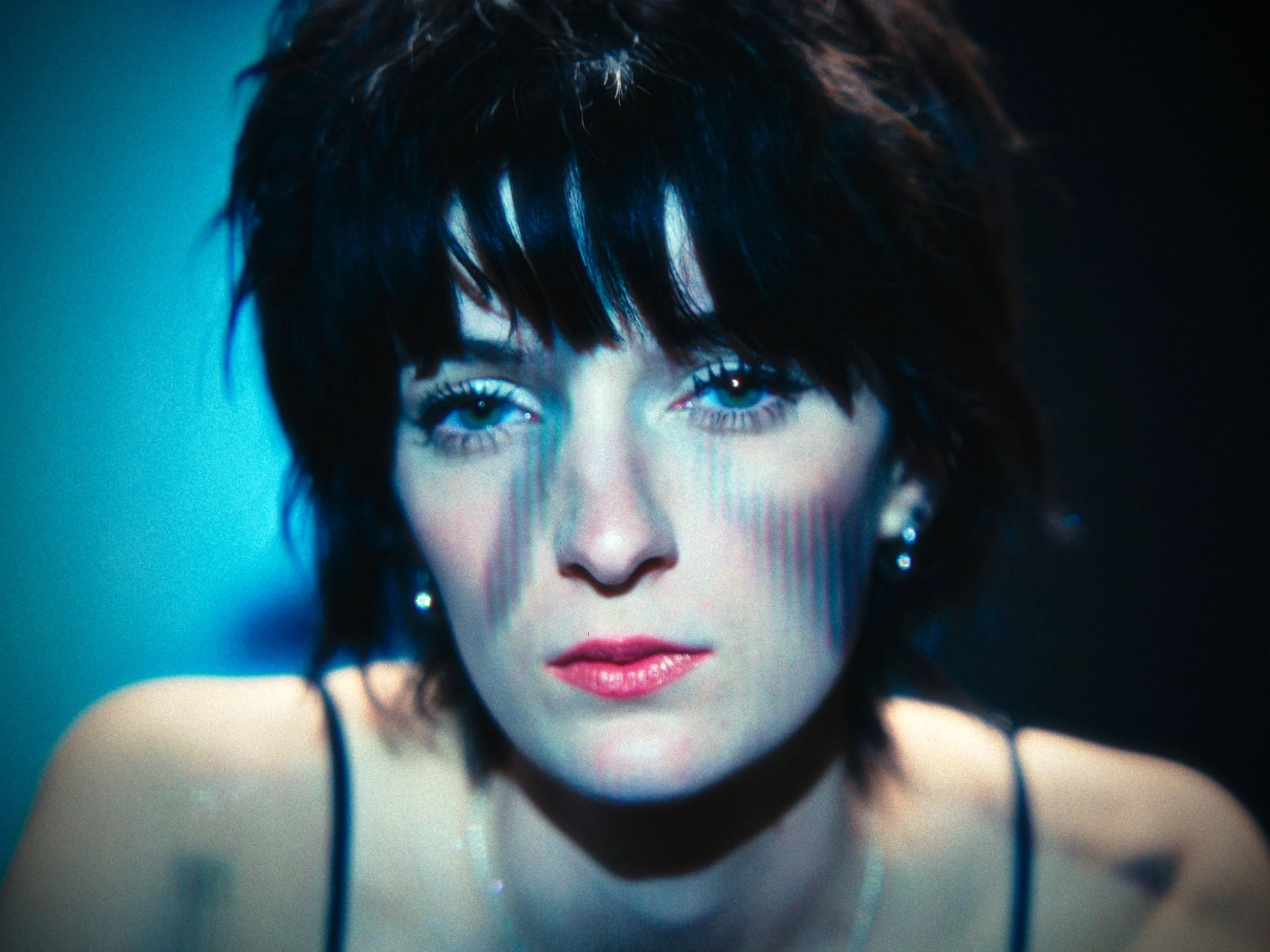 Close-up of a woman with short dark hair, striking blue eyes, and makeup, wearing earrings and a thin-strapped top, with a dark background.