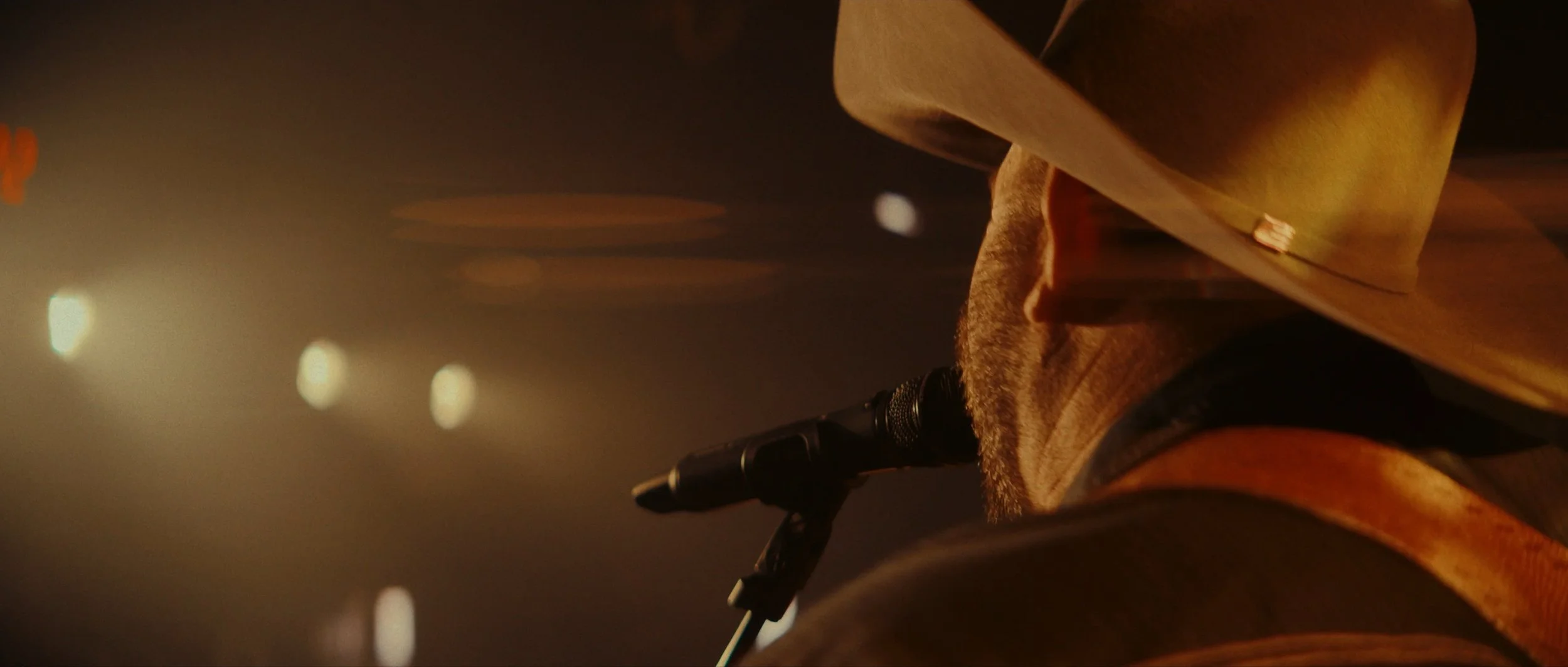 A man wearing a cowboy hat singing into a microphone during a performance, with stage lights in the background.