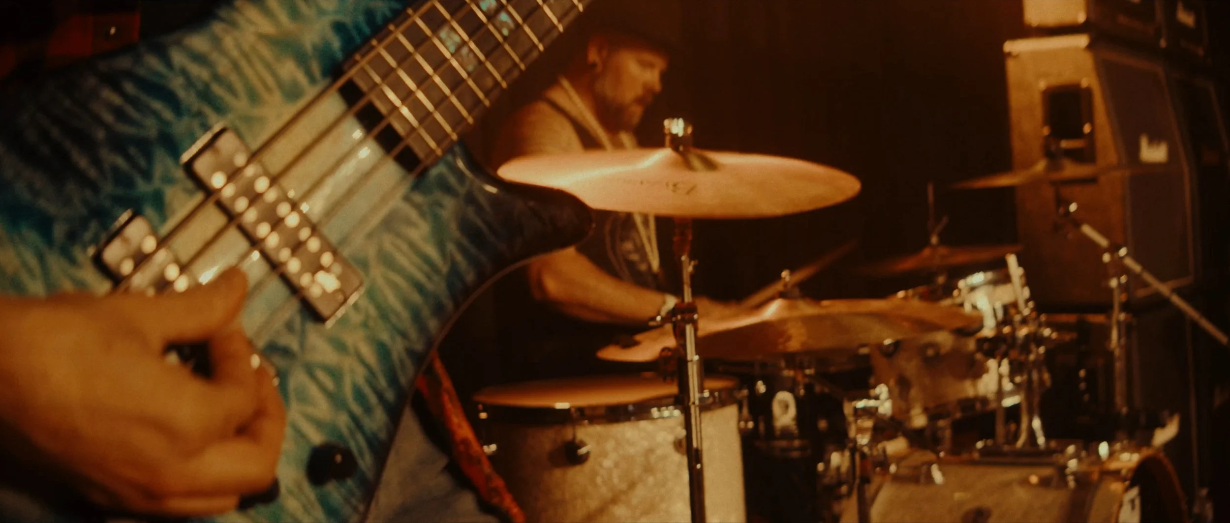 Close-up of an electric guitar neck with tuning pegs and fretboard in the foreground, and a drummer playing on stage in the background with drums and speakers, performing in a dimly lit setting.