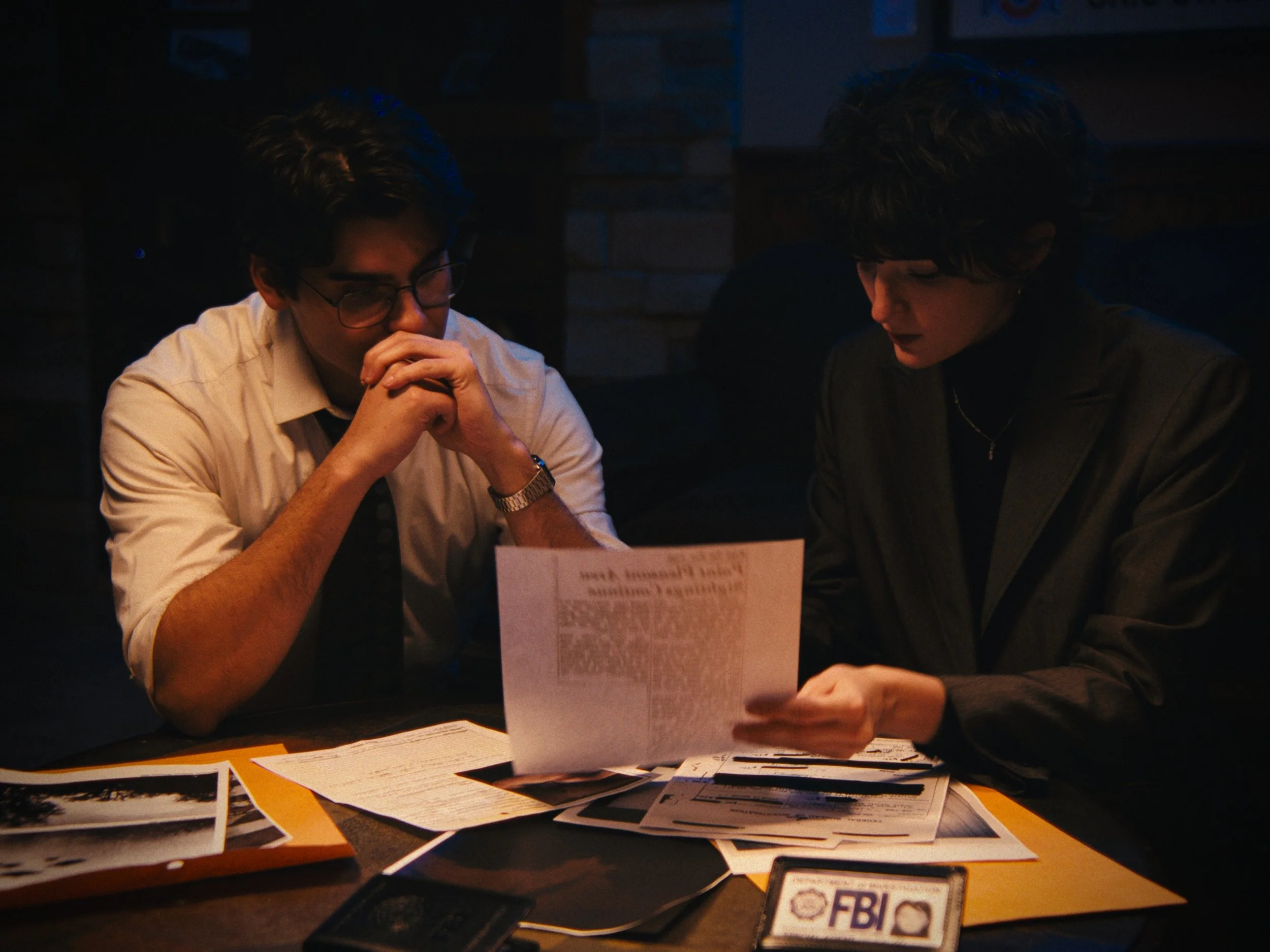 Two individuals, a man and a woman, sit at a table examining documents under dim lighting. The table has various papers and an FBI badge visible.