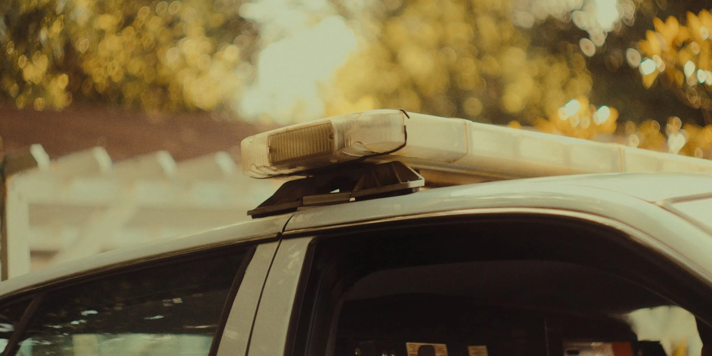 Close-up of police vehicle emergency light bar on top of a car, with a blurred background of trees and structures.