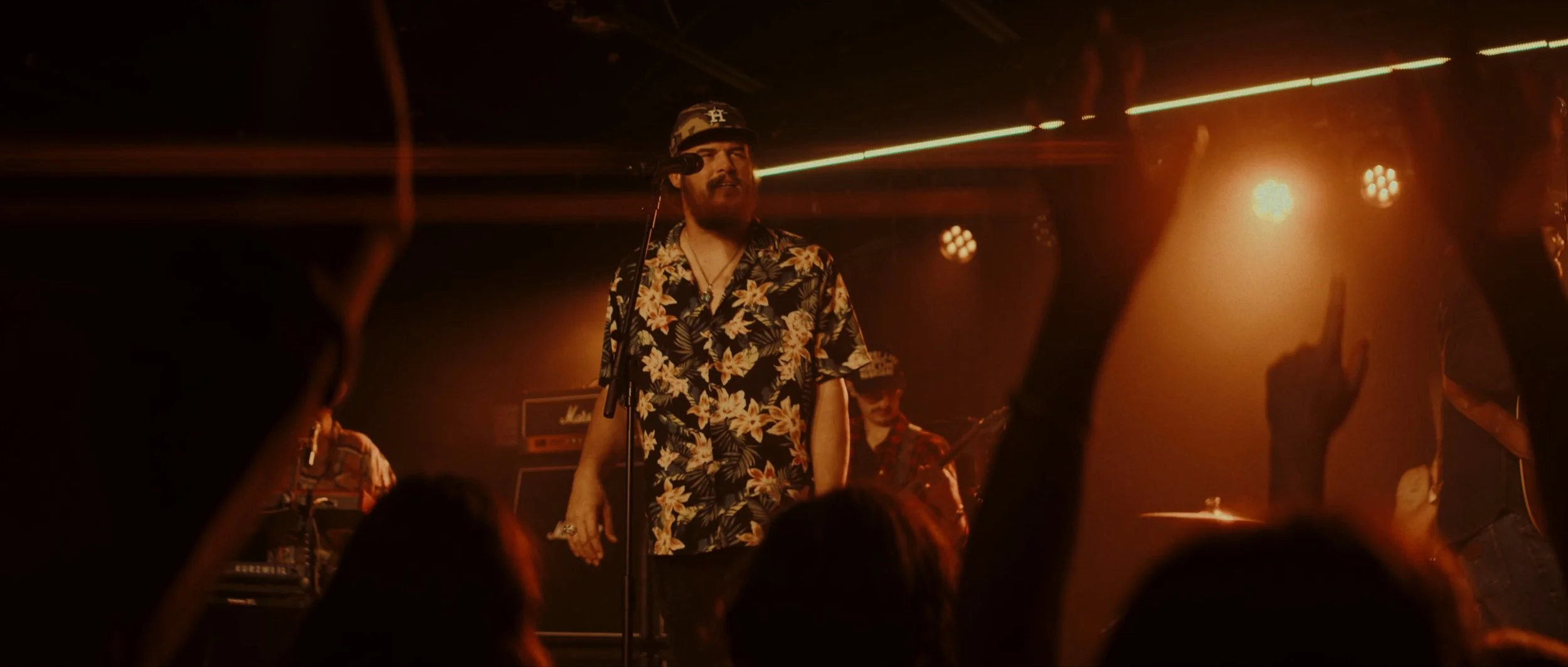 A man with a beard wearing a floral shirt and a baseball cap, standing on stage with a microphone, performing at a concert with orange lighting. Audience hands are raised in the foreground.