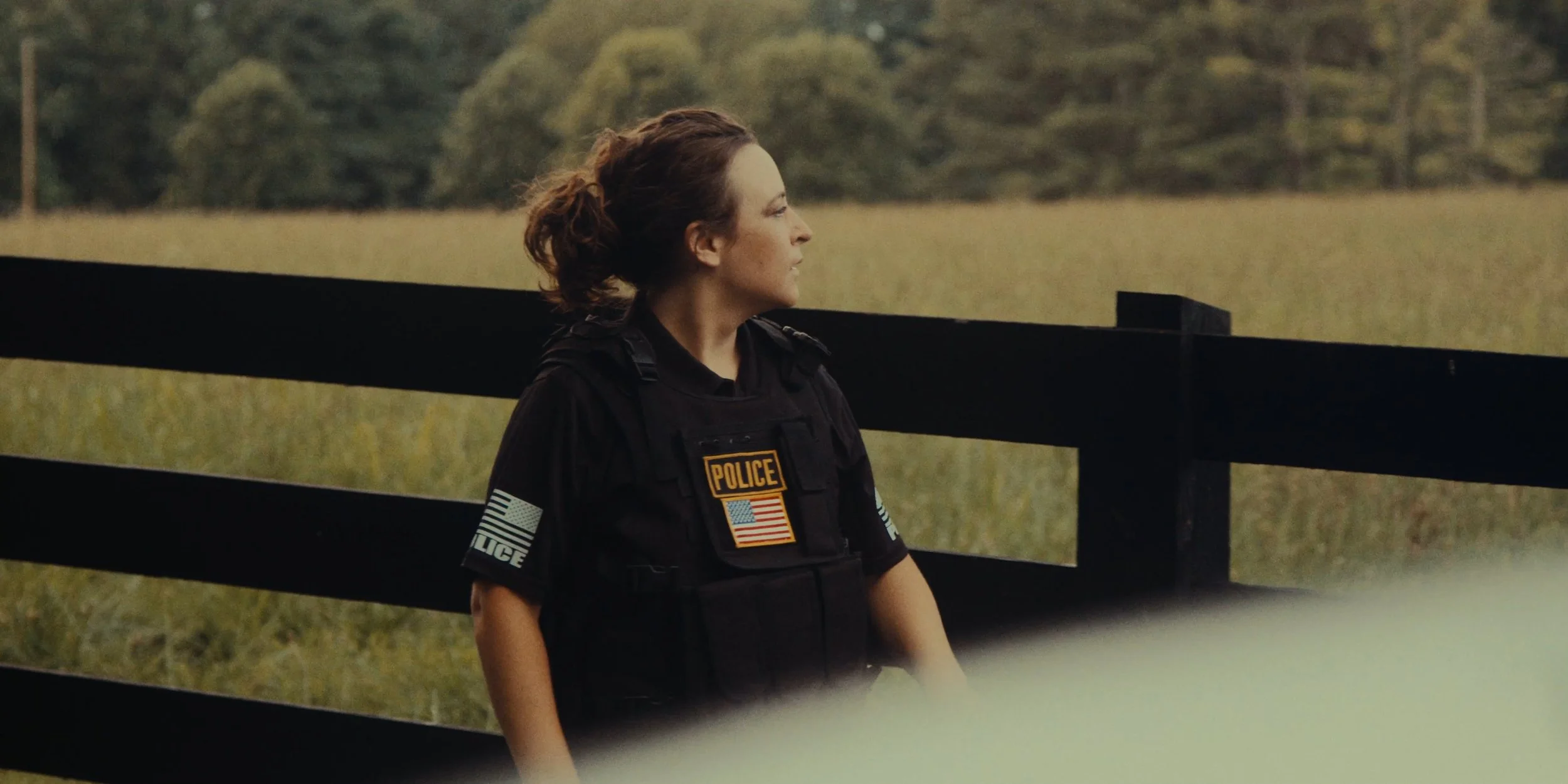 A female police officer standing outdoors in front of a black wooden fence and grassy field, looking to the side.