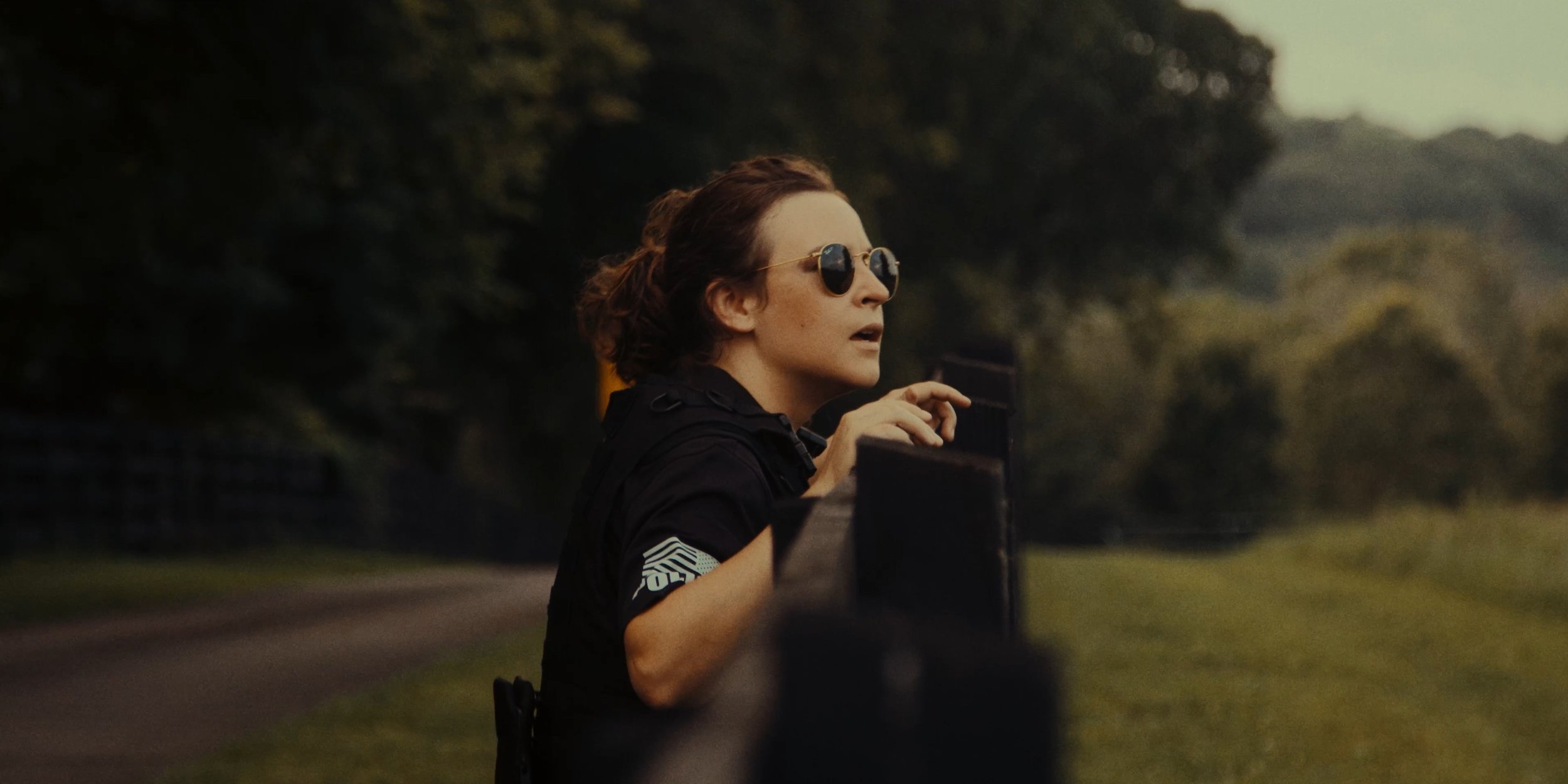 A woman with short brown hair and sunglasses pointing as she looks into the distance outdoors.