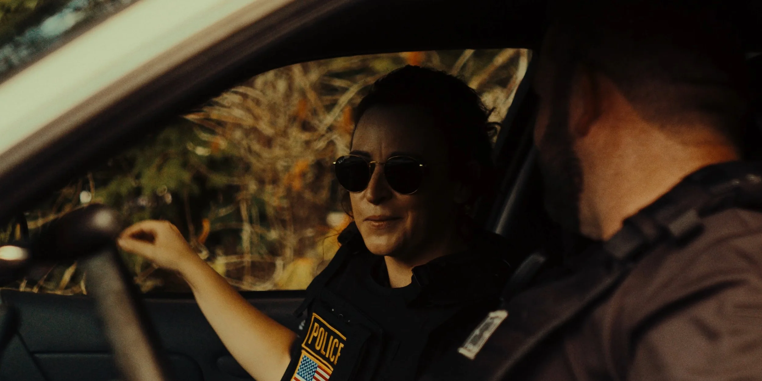 A female police officer and a male police officer inside a patrol car, with the female officer wearing sunglasses and a police uniform, sitting in the driver's seat.