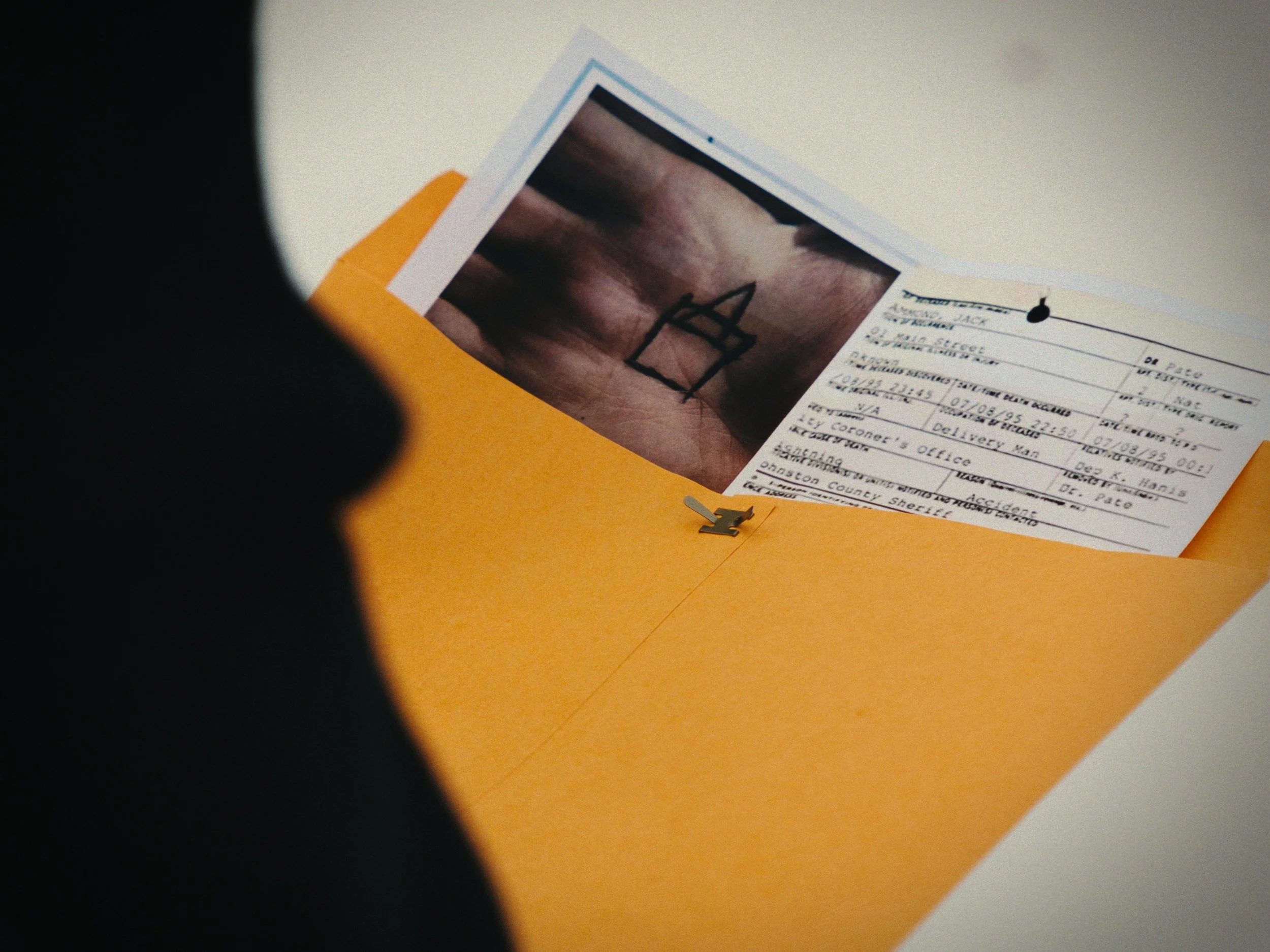 A partially opened orange envelope revealing a photograph and a form inside. The photograph appears to be a close-up of a hand. The form contains typed text and handwritten notes and is secured with a black pushpin.
