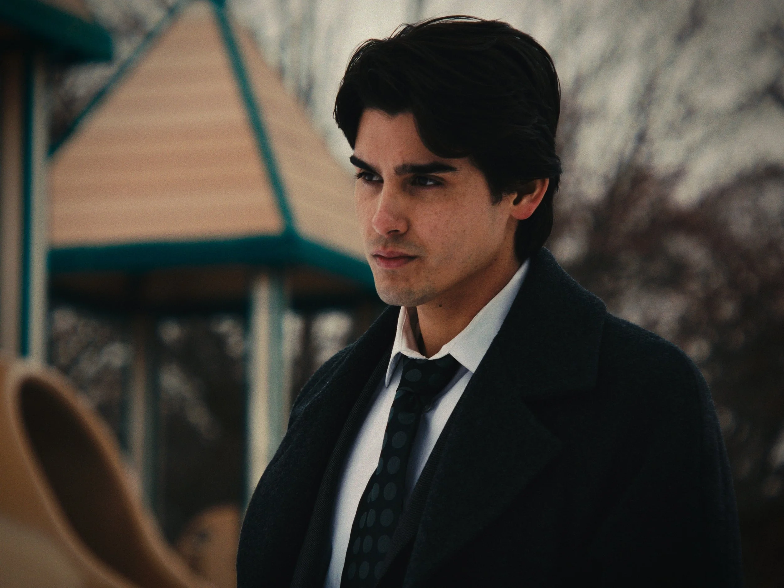 A young man with dark hair, wearing a dark coat, white shirt, and a dark polka dot tie, standing outdoors with a blurred playground in the background.
