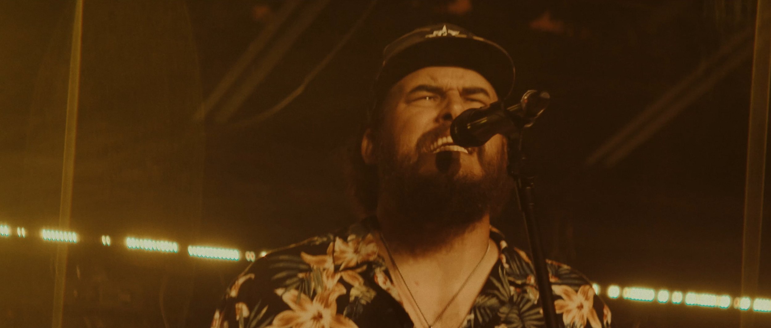 A man with a beard and mustache singing into a microphone on stage, wearing a floral shirt and a cap, with stage lights in the background.
