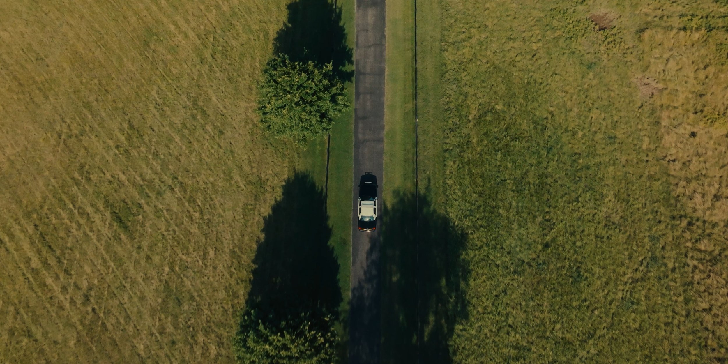 An aerial view of a white and black vehicle on a narrow paved road flanked by trees and grassy fields.