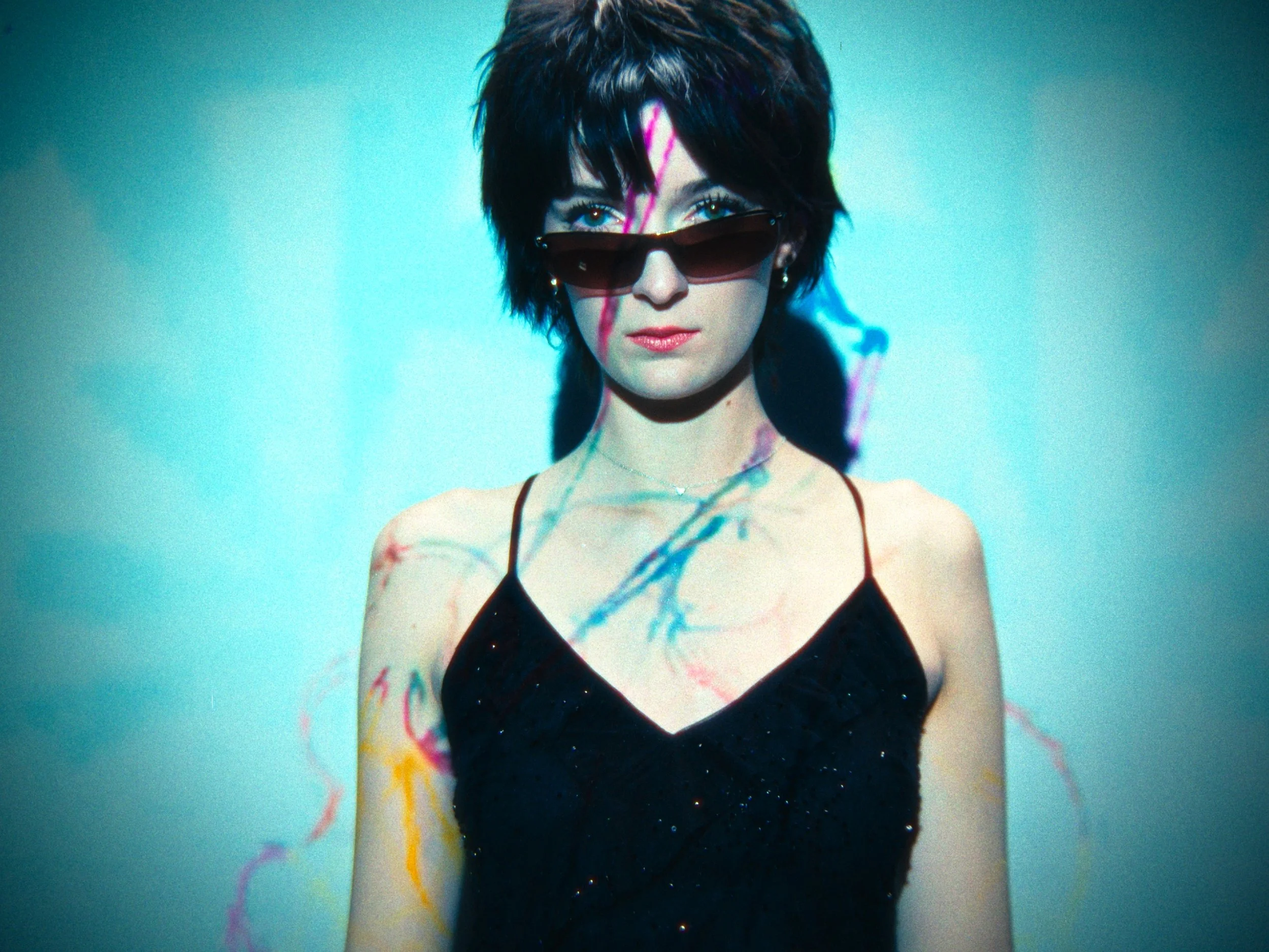 A young woman with short dark hair, wearing sunglasses and a black dress, standing against a brightly lit background with colorful painted lines on her skin.
