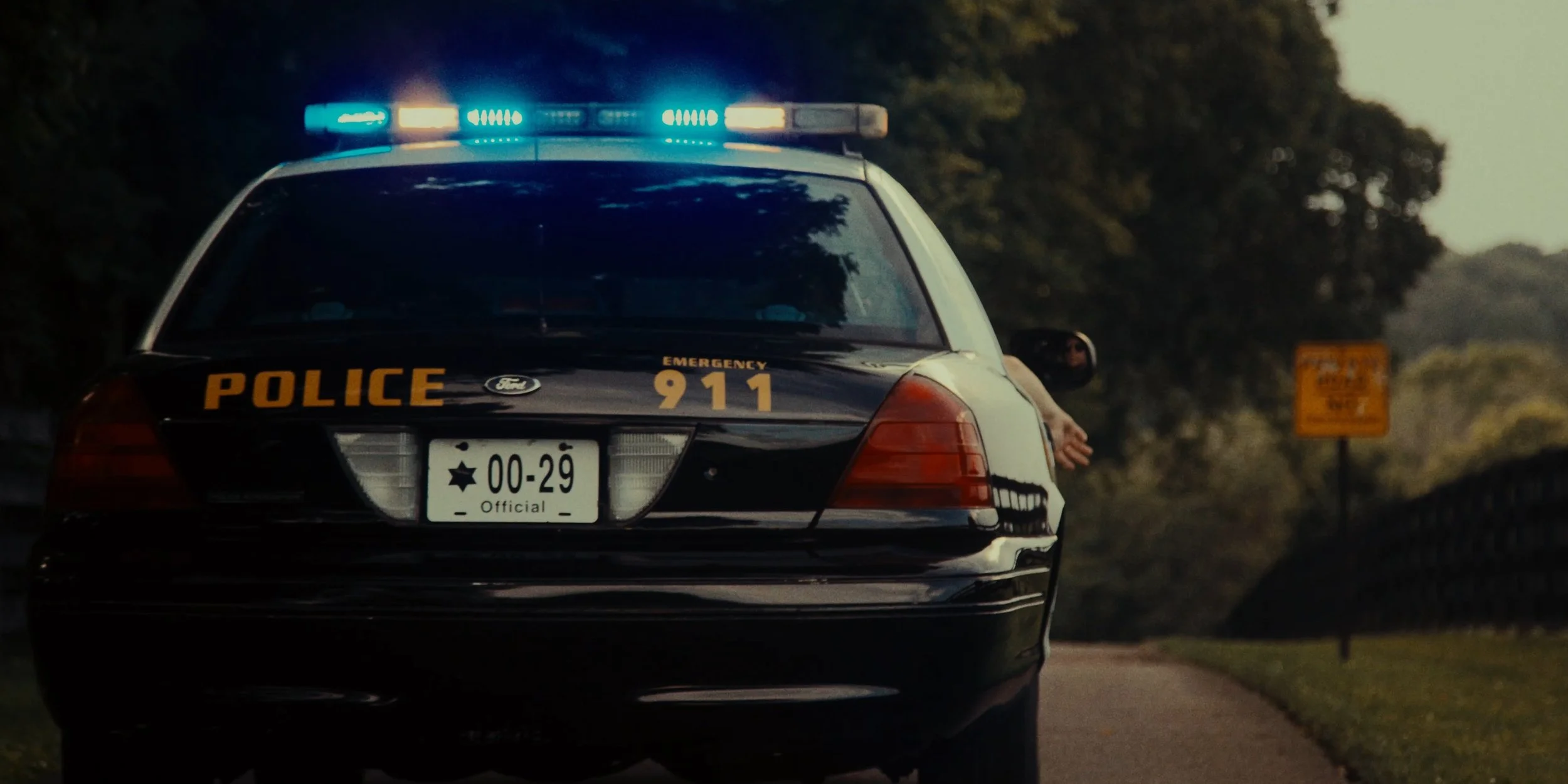 A police car with flashing blue lights on a rural road at dusk, with a person reaching out of the window and a yellow warning sign in the background.