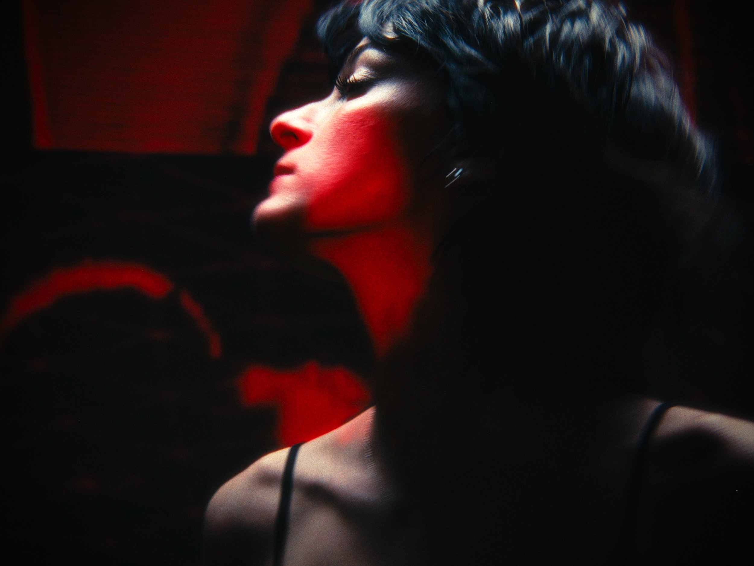 Close-up of a woman's face in profile with dramatic lighting, dark hair, and red lipstick, with a blurred dark background.