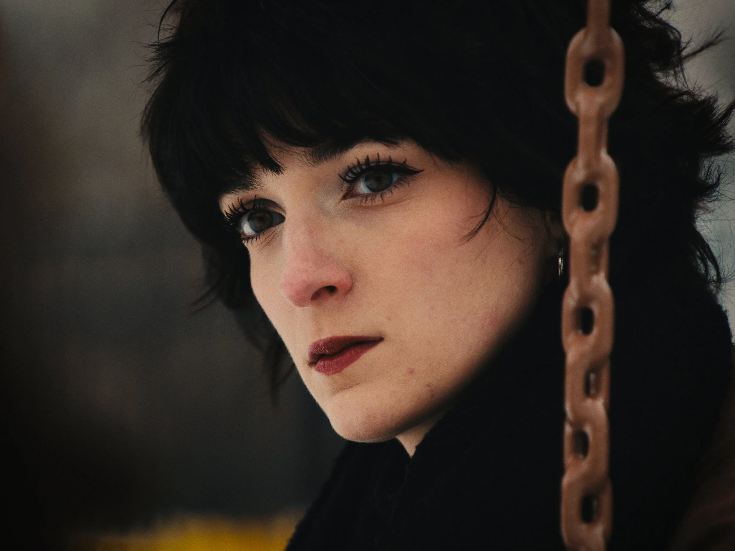Close-up of a woman with short black hair, fair skin, and dramatic makeup, partially obscured by a hanging chain on the right side of the image.