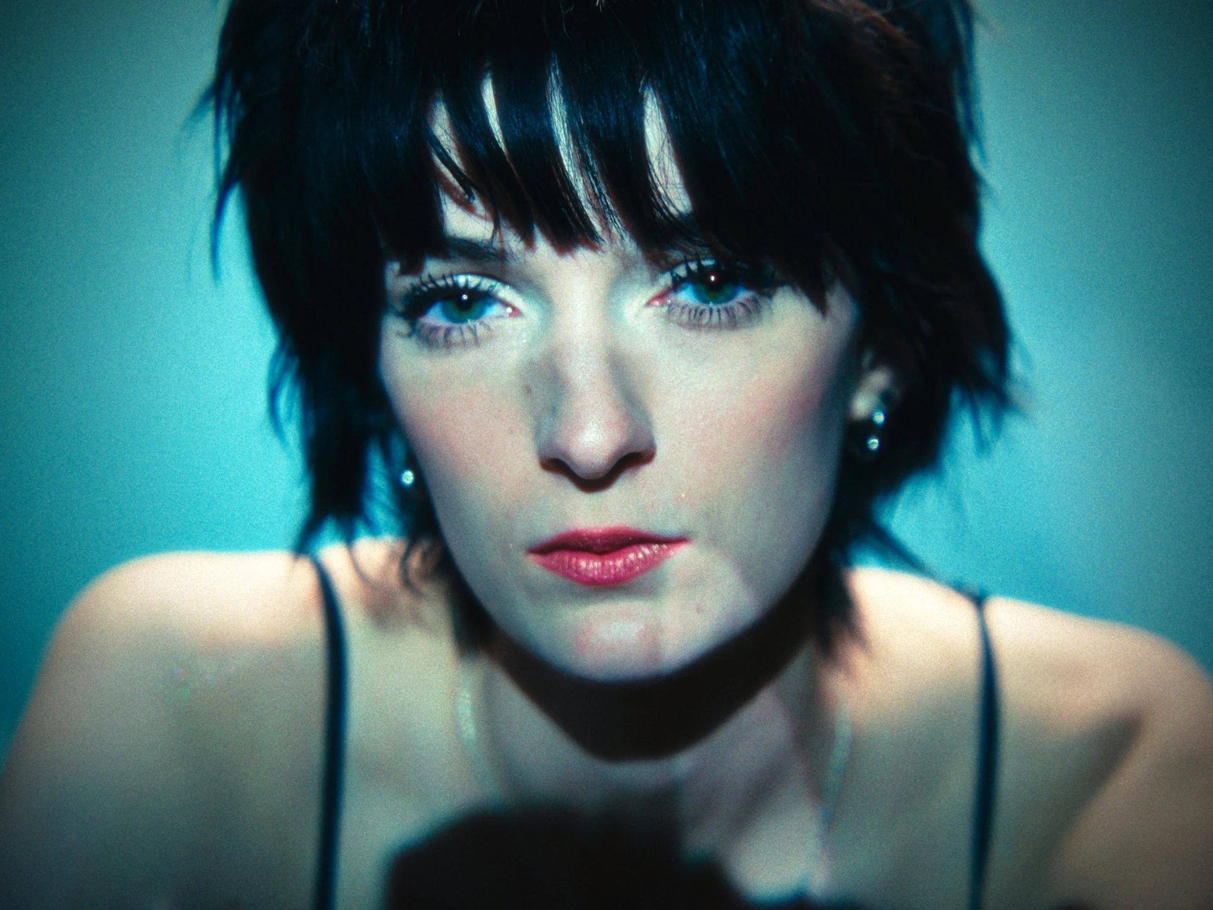 Close-up of a woman with short black hair, blue eyes, and wearing earrings, looking directly at the camera against a blue background.
