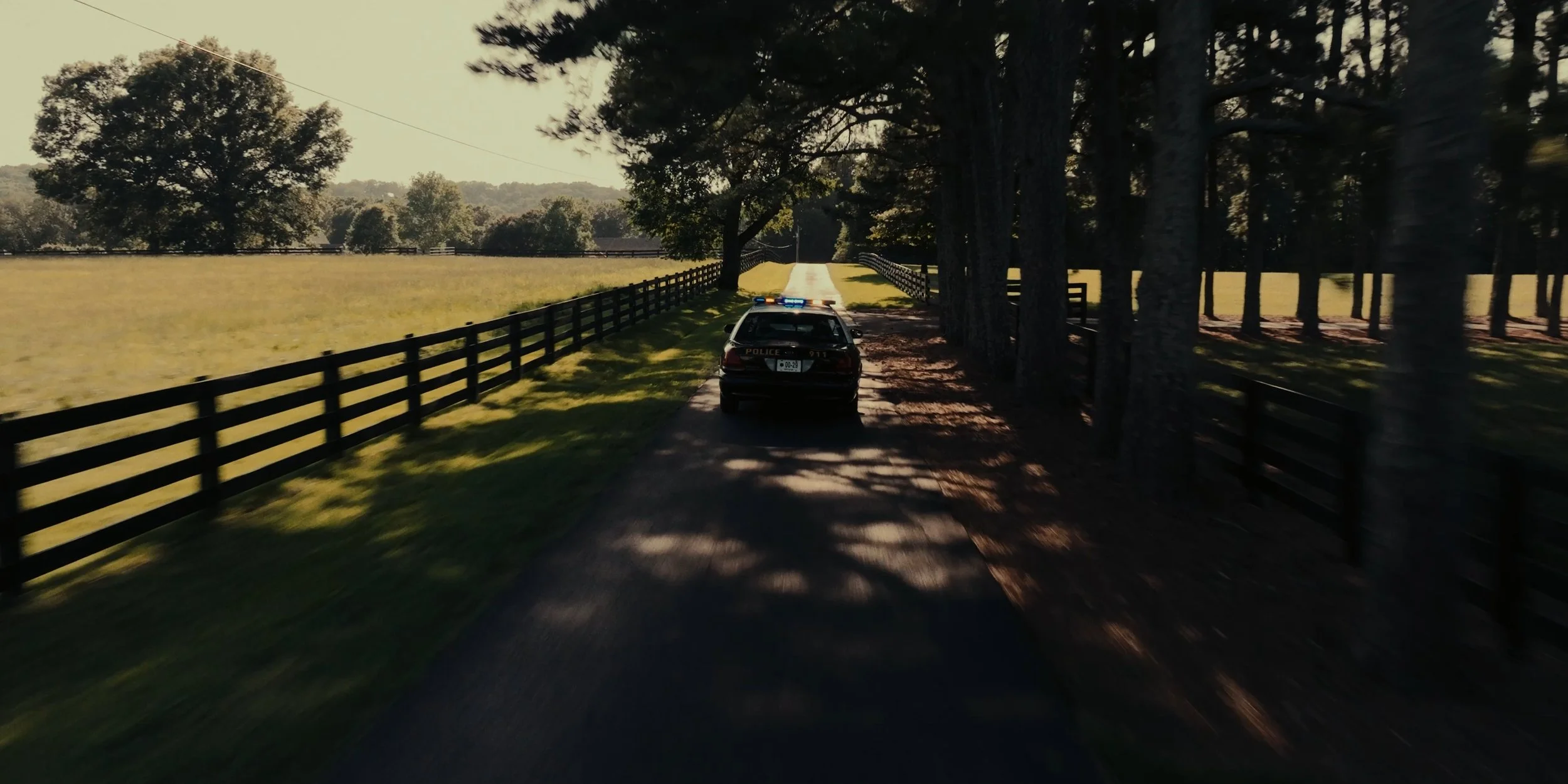A police car with flashing lights parked on a rural dirt road surrounded by fencing, trees, and open fields on a sunny day.